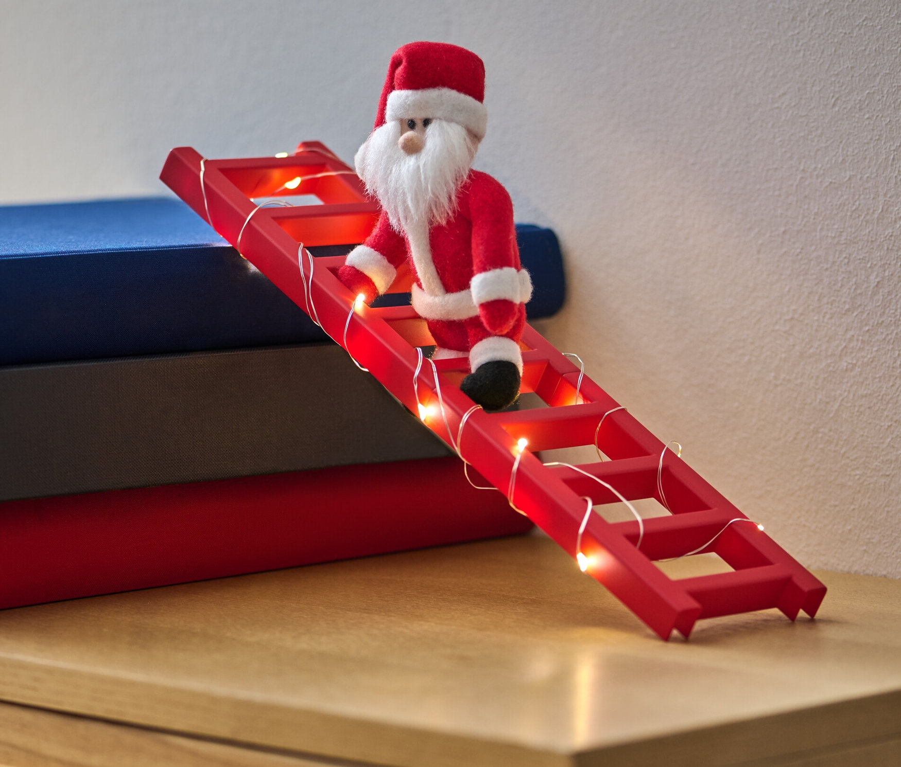 Eine Santa Claus Puppe klettert auf eine rote Leiter mit LED-Lichterkette, die an einen Bücherstapel gelehnt ist.