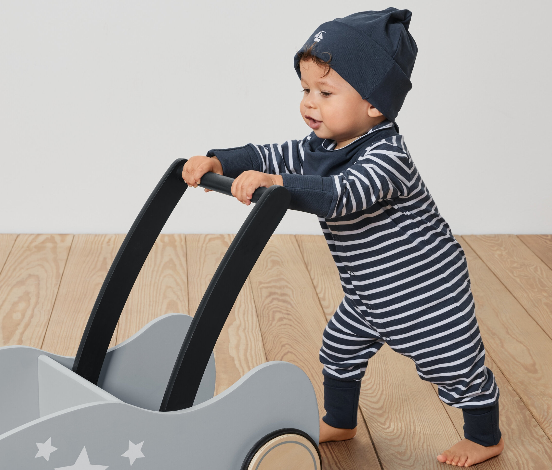 Kleinkind schiebt grauen Wagen mit Sternen. Es trägt ein Baby-Set, 3-teilig: gestreifter Overall, Mütze und Lätzchen.