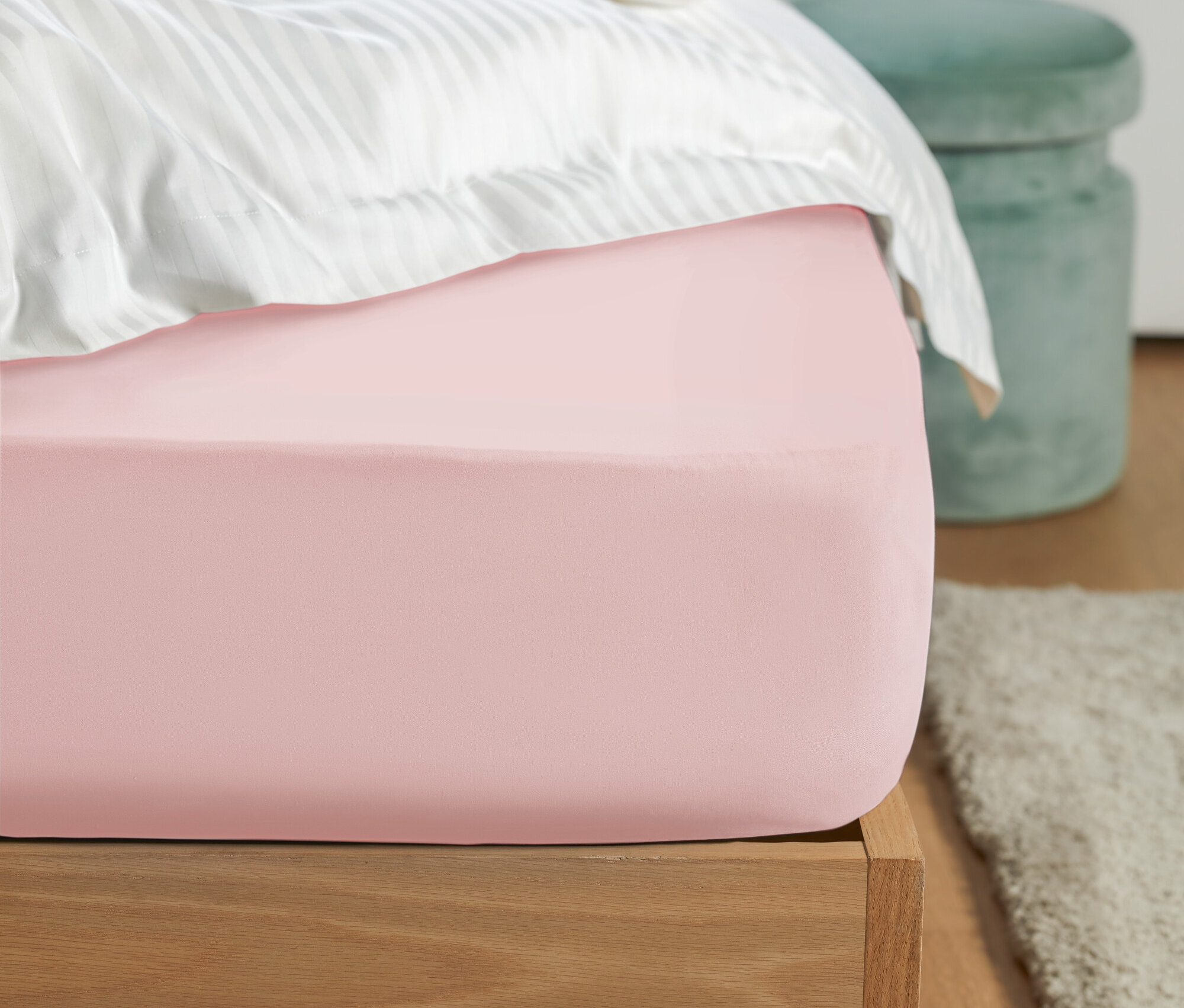 Detailaufnahme eines rosa Spannbetttuchs auf einem Bett mit weiß gestreifter Bettwäsche und einem grünen Hocker im Hintergrund.