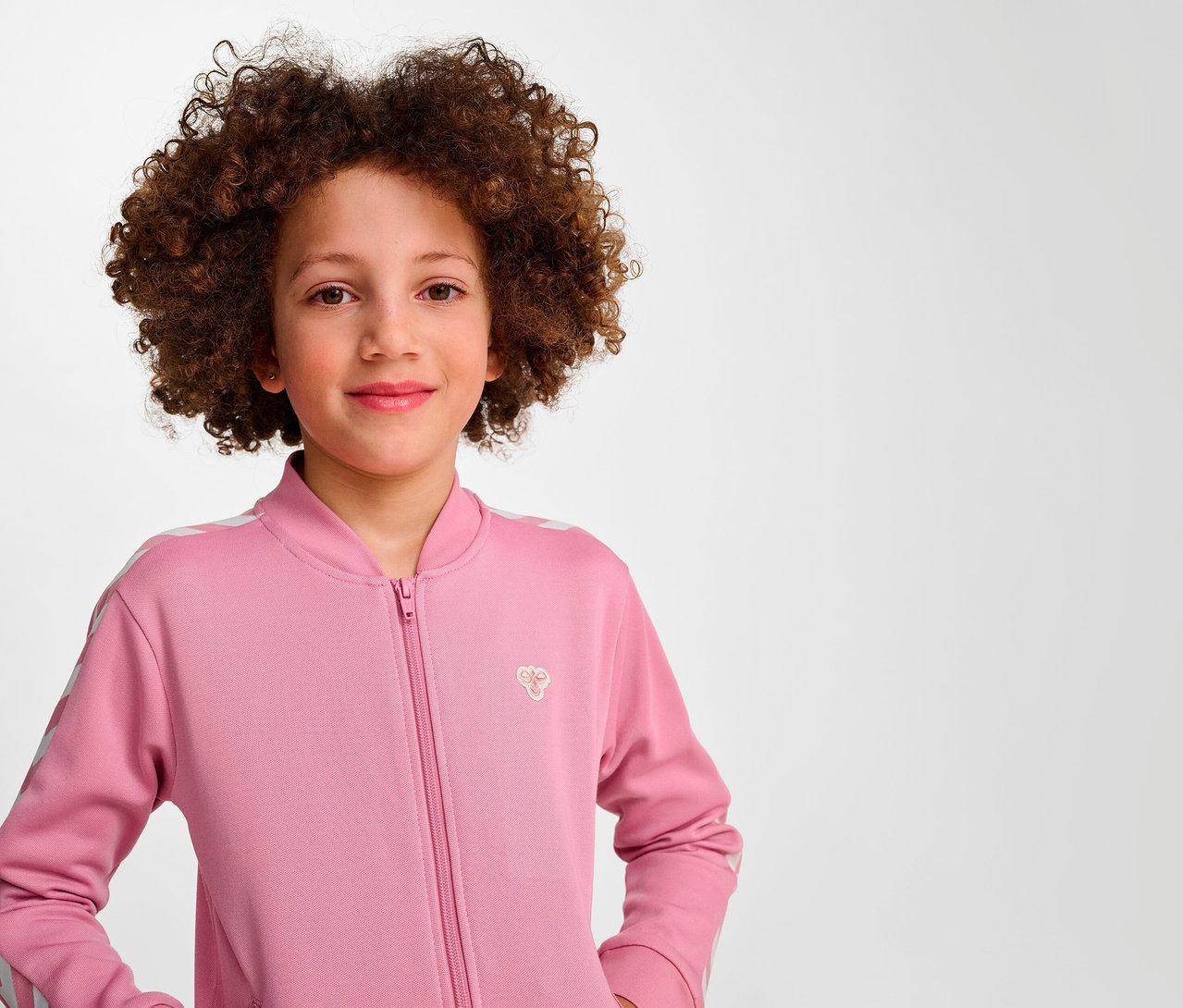 Porträt eines lächelnden Mädchens mit Afro in einer rosa Hummel HMLJR Archive Reg Poly Zip Jacket.