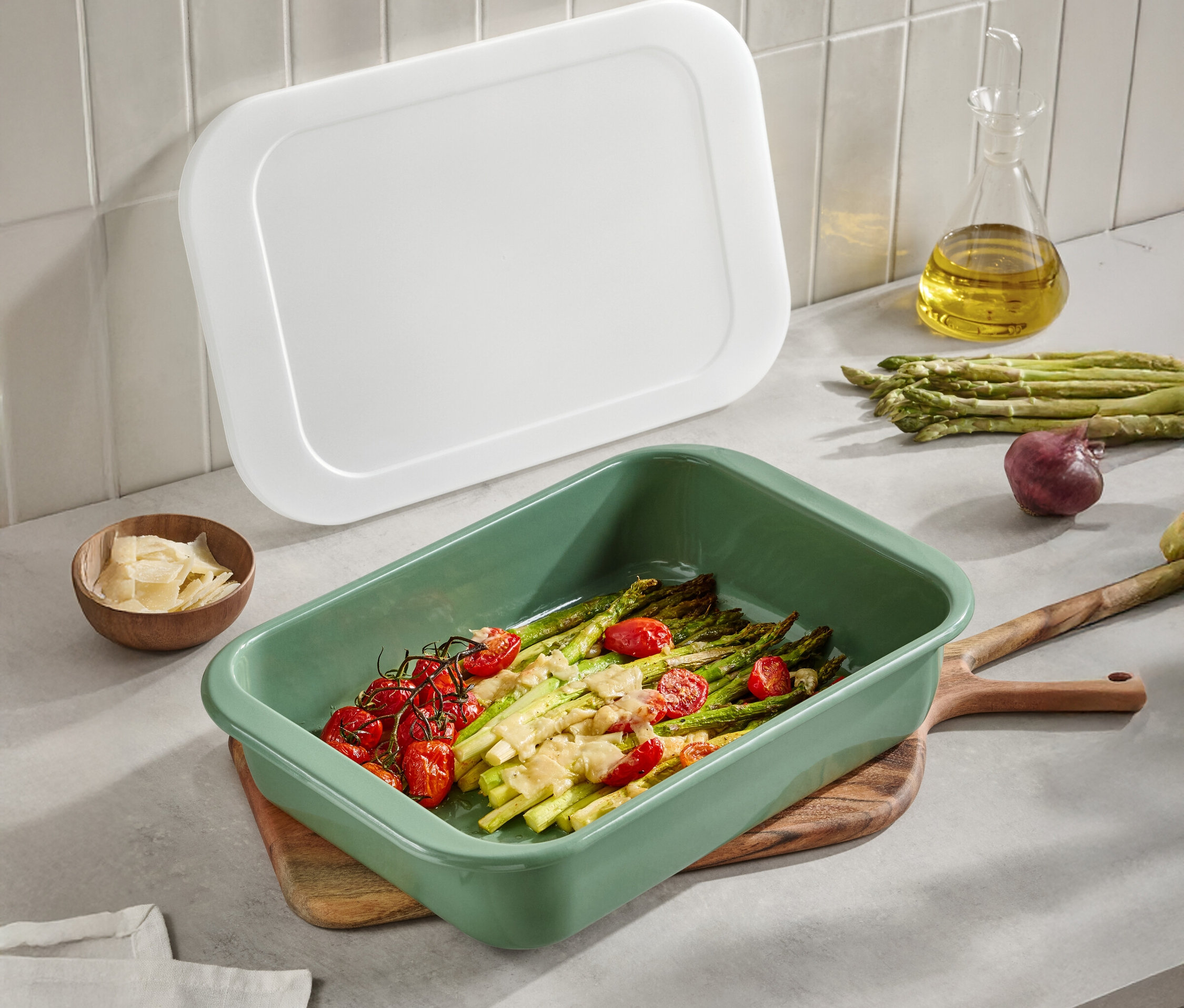 Grüne Auflaufform mit Spargel und Tomaten auf einem Holztisch. Eine weiße Abdeckung, Olivenöl, Parmesan und Zwiebeln sind ebenfalls sichtbar.