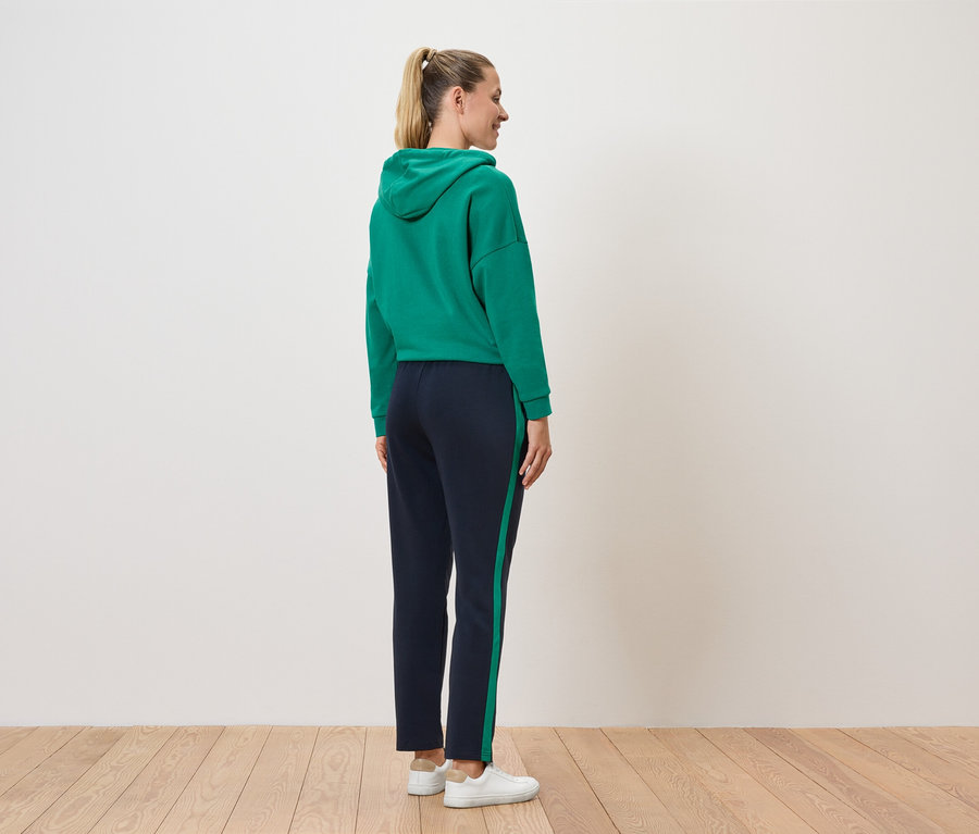 Frau steht mit dem Rücken zur Kamera und trägt einen grünen Kapuzenpullover und eine dunkelblaue Hose mit Streifen.