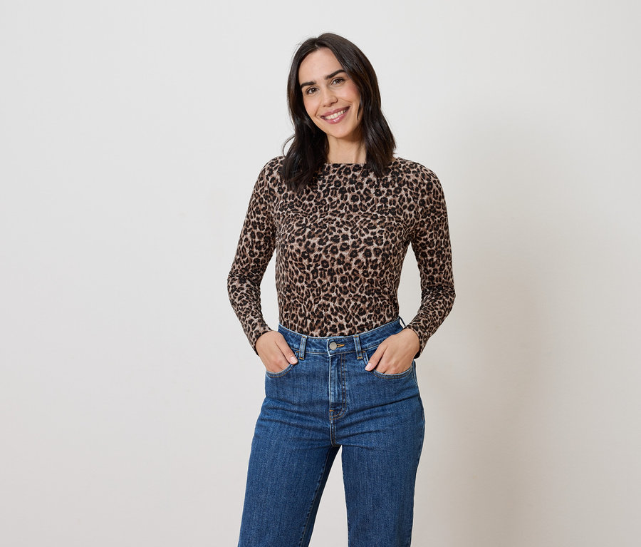 Frau mit braunem Haar posiert in einem Oberteil mit Tiermuster und blauen Jeans.