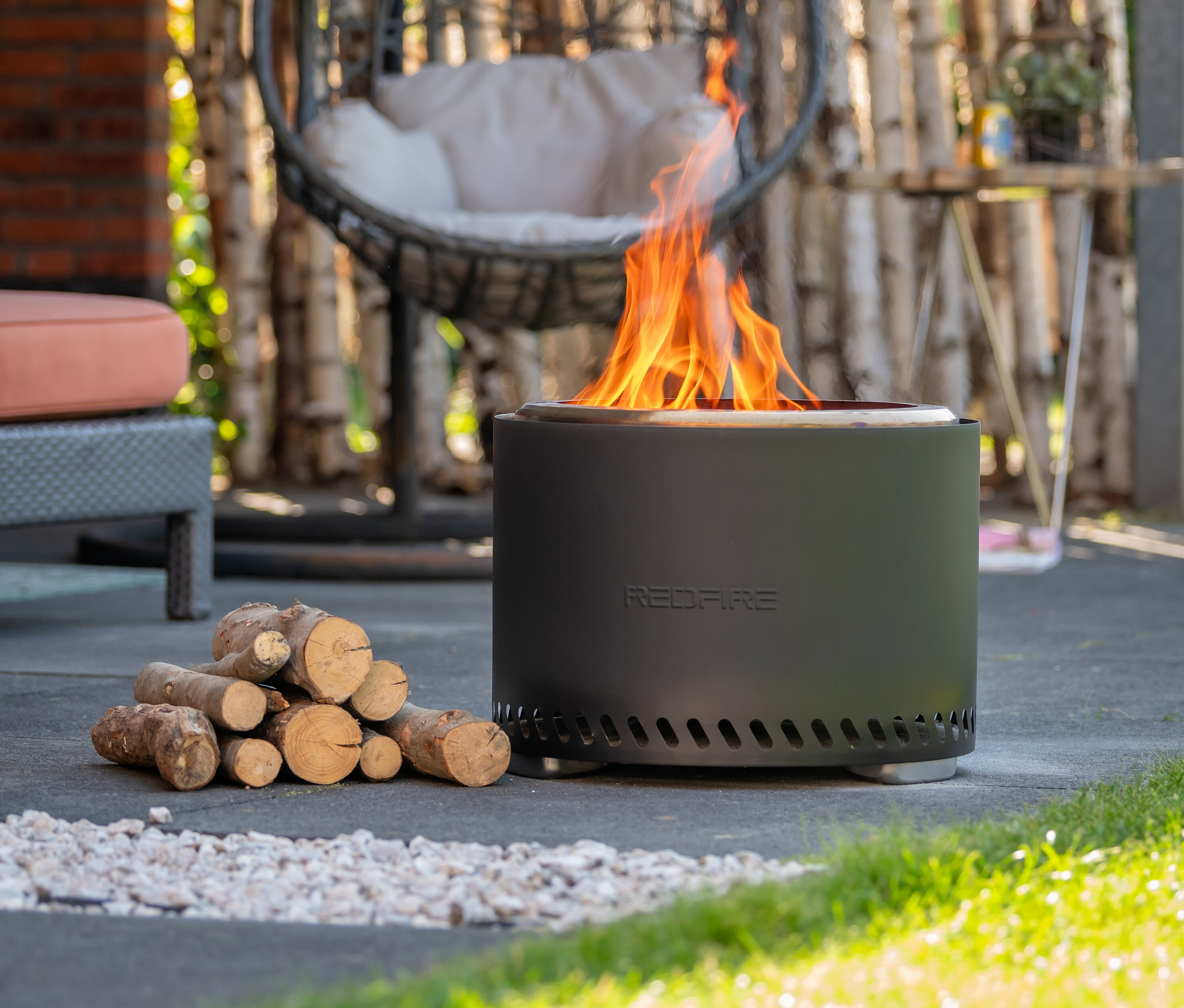 Eine RedFire Feuerschale »Volcano Extra Large« mit Flammen und einem Holzstapel daneben.