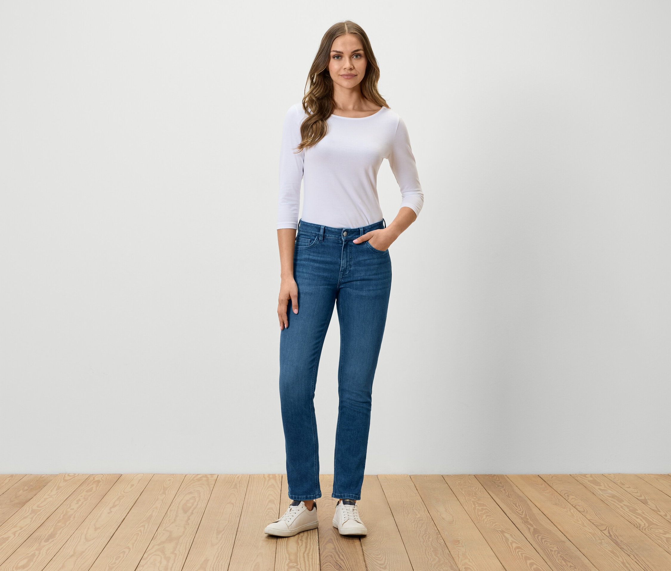 Frau steht und posiert in blauen Jeans, weißem T-Shirt und weißen Turnschuhen.