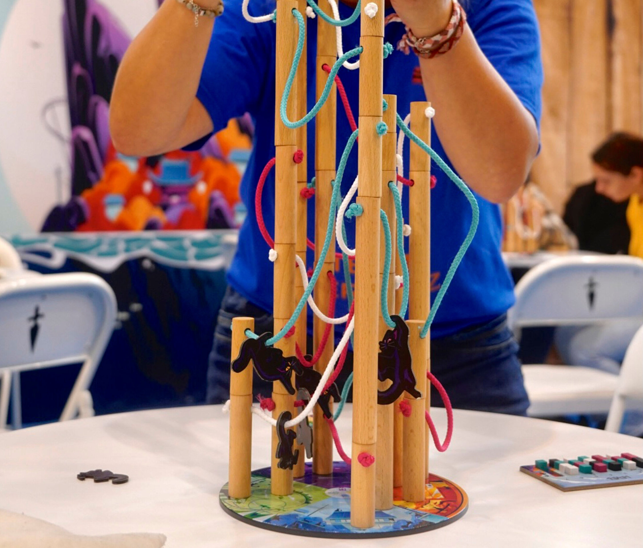 Eine Person spielt mit dem »Nekojima« Spiel, das aus Holzstäben, Schnüren und Katzenfiguren besteht.