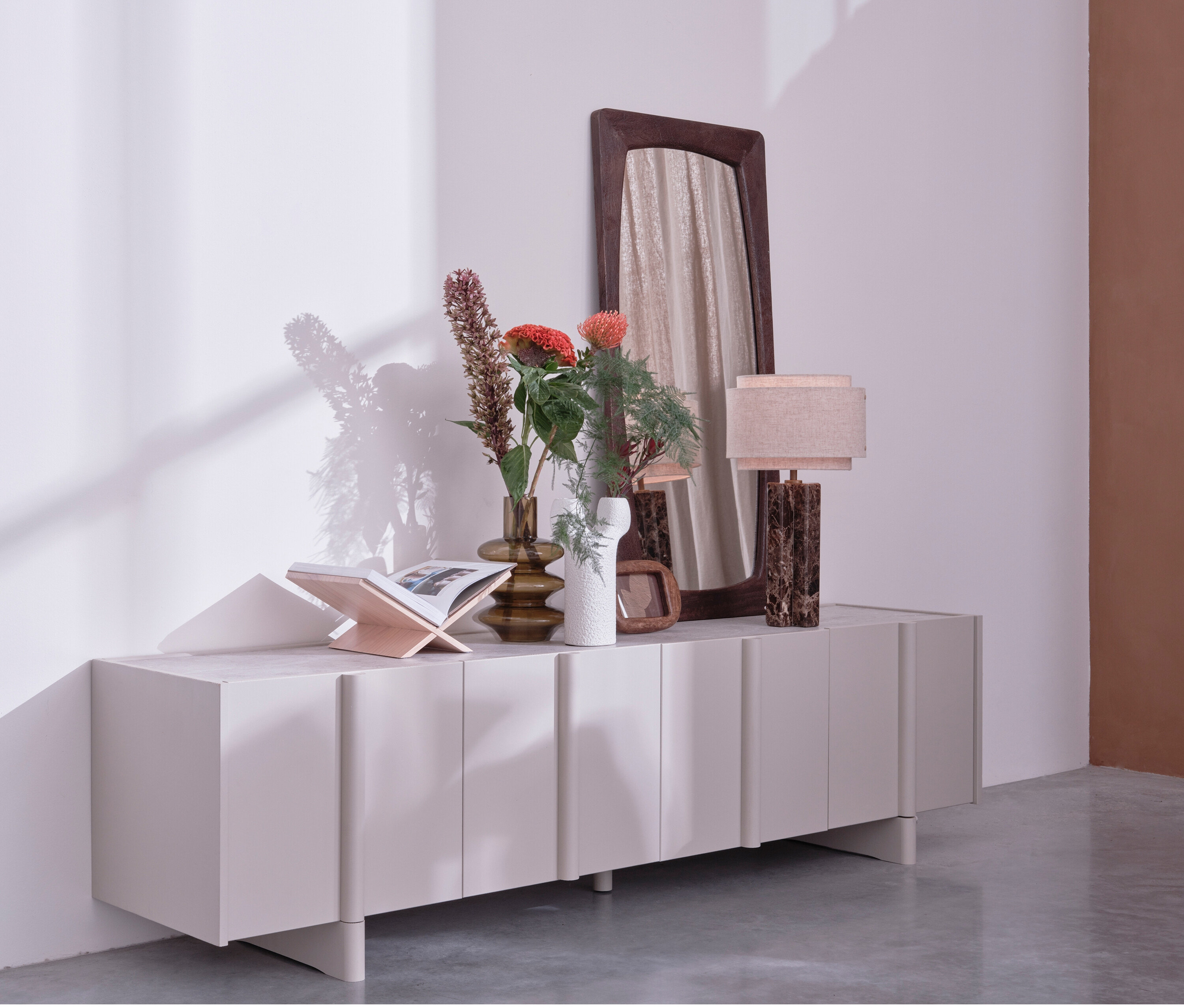 Auf einem WOOOD Lowboard »Basu« kiefer dust stehen Vasen mit Blumen, ein Buch, eine Lampe und ein Spiegel.