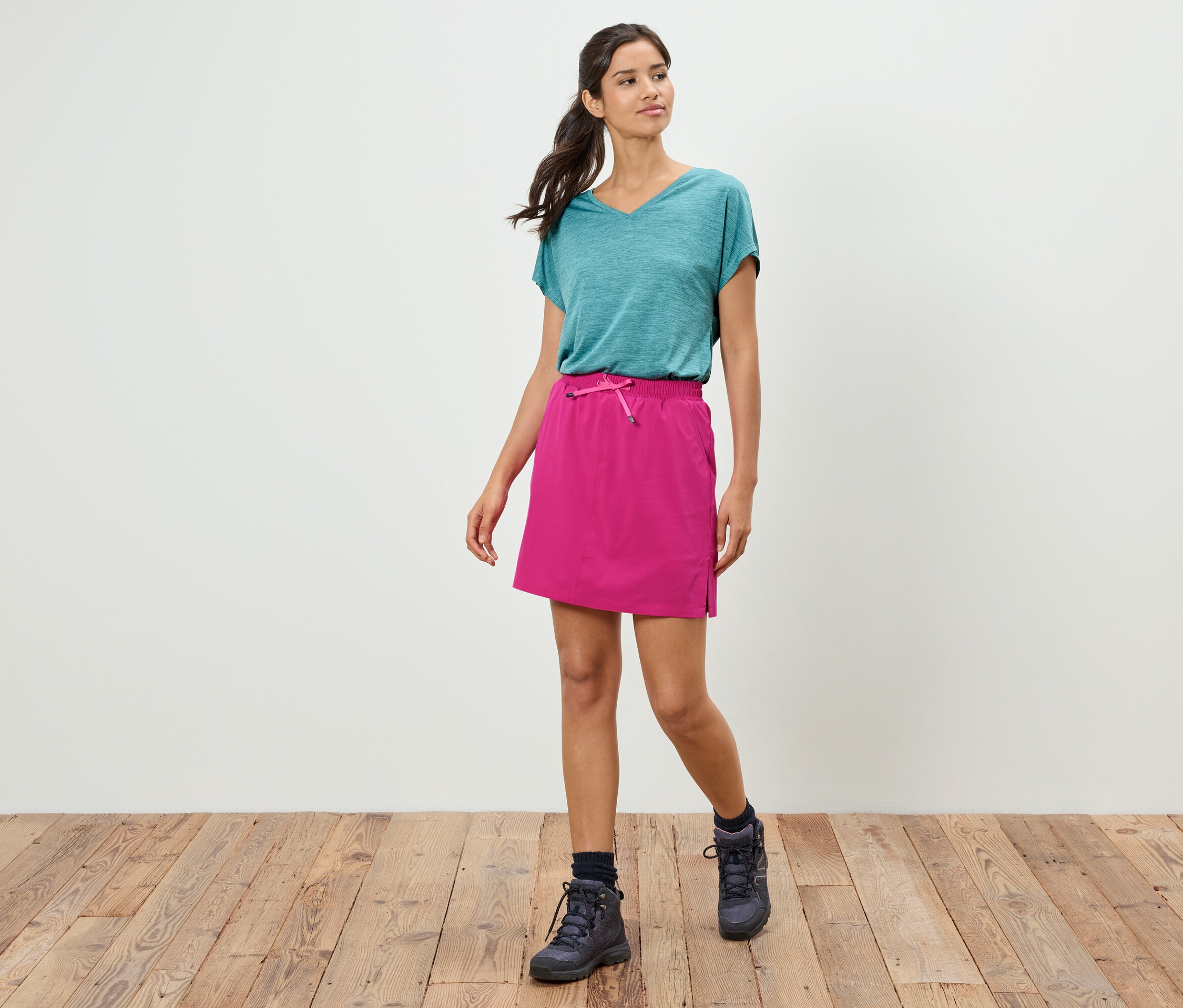 Frau geht in blaugrünem T-Shirt, pinkfarbenem Rock und Wanderschuhen auf Holzboden vor einer weißen Wand.