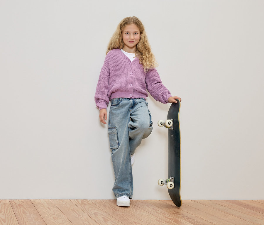 Mädchen mit blonden Locken, trägt einen lila Strickjacke und Jeans, lehnt an einem Skateboard vor einer weißen Wand.