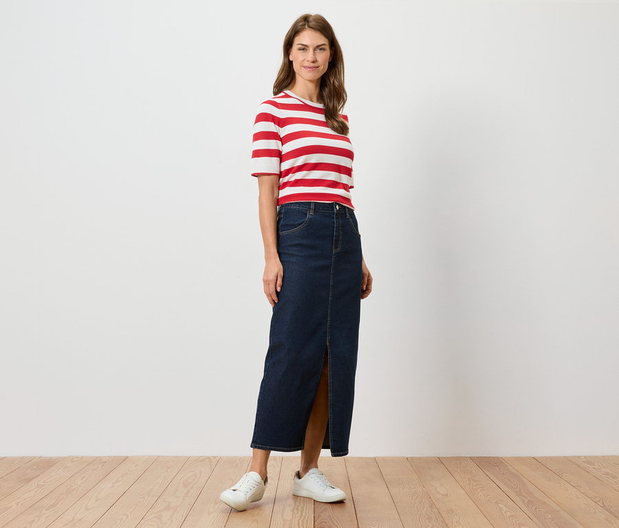 Eine Frau steht und trägt ein rot-weiß gestreiftes T-Shirt, einen langen Jeansrock mit Schlitz und weiße Turnschuhe.