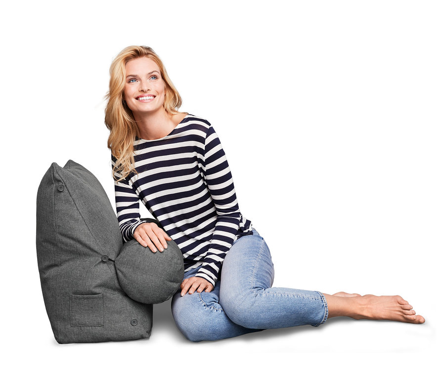 Lächelnde Frau sitzt neben einem grauen Rückenkissen mit Nackenrolle.