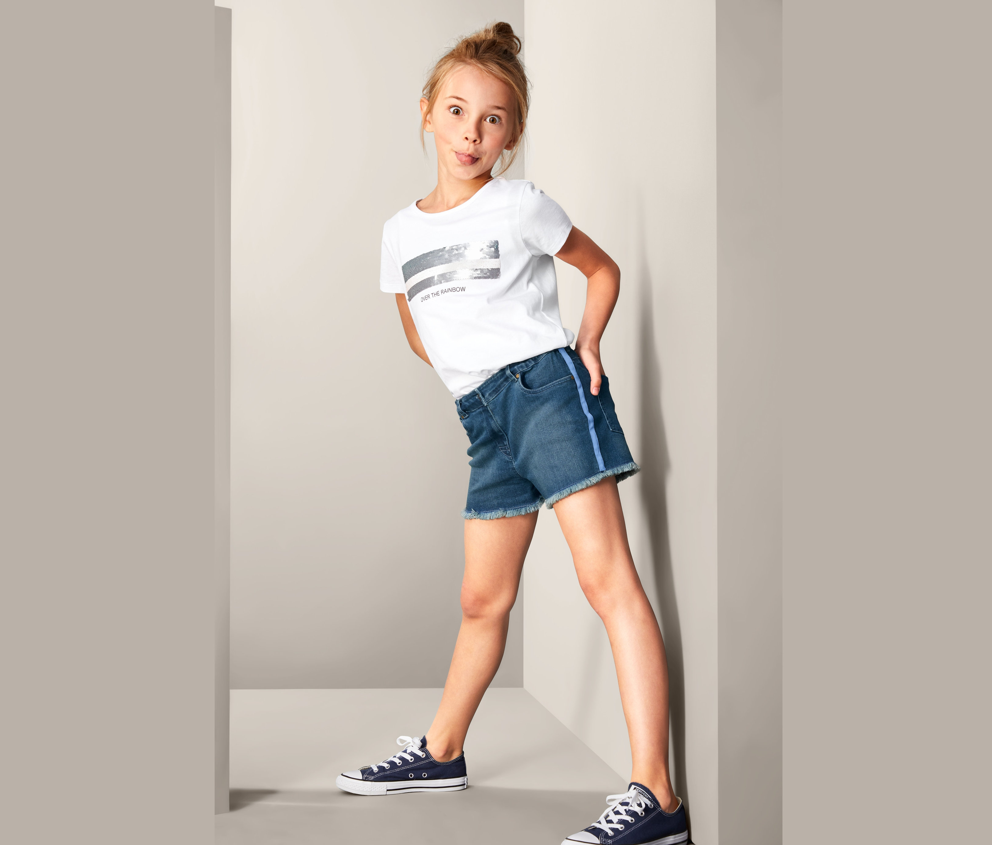 Mädchen posiert in Denim-Shorts, T-Shirt und Turnschuhen.