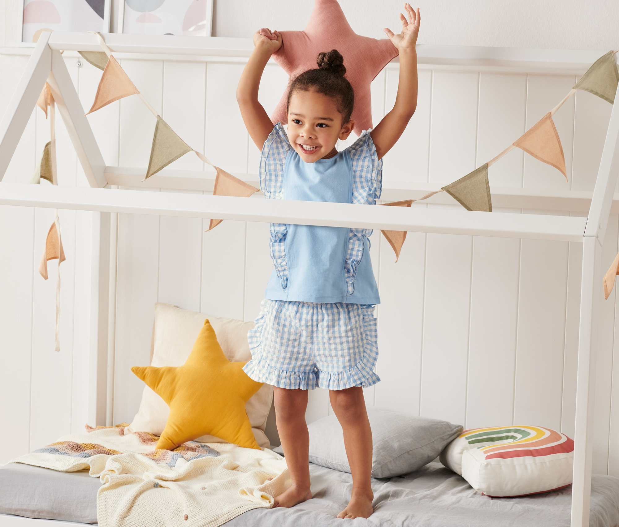 Ein Mädchen steht in einem Hausbett und hält einen rosa Stern über den Kopf. Sie trägt eine blau-weiß karierte Vichy-Karo-Webshorts aus Bio-Baumwolle.