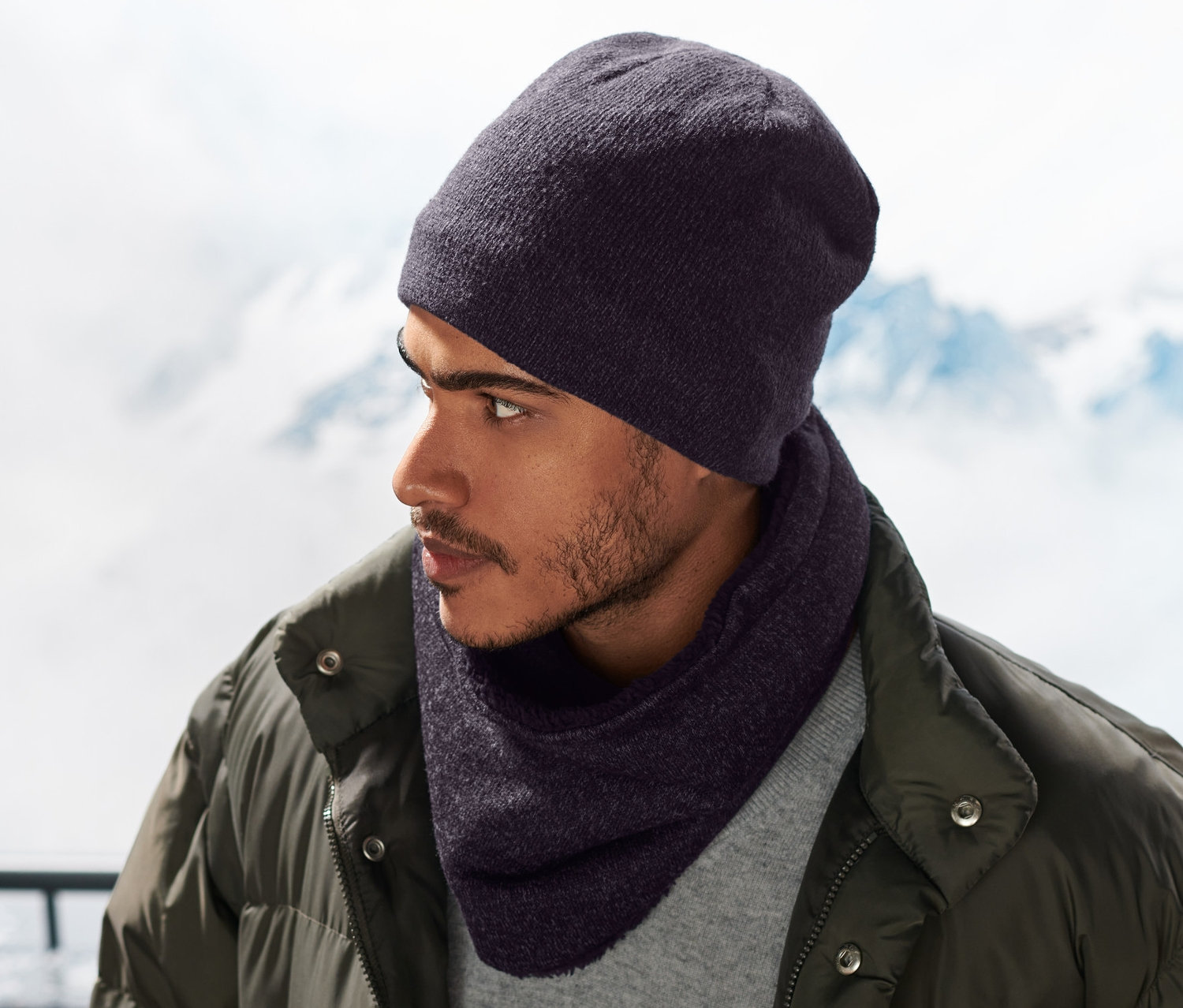 Mann trägt einen blauen Strickfleece-Schlauchschal und eine Mütze, im Hintergrund schneebedeckte Berge.