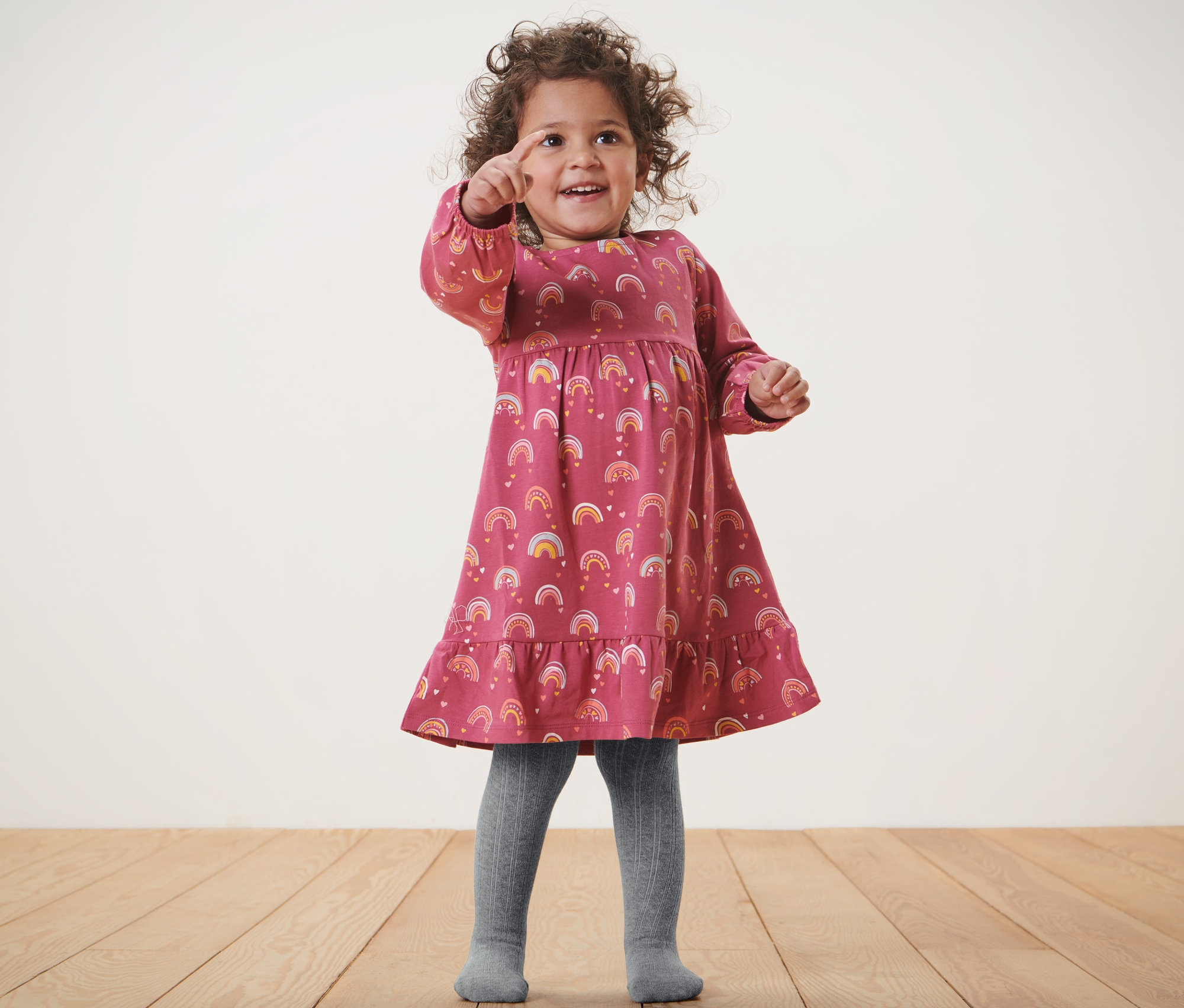 Mädchen im Kleid mit Regenbogenmuster und grauer Baby-Strumpfhose, 2 Paar, zeigt mit dem Finger.