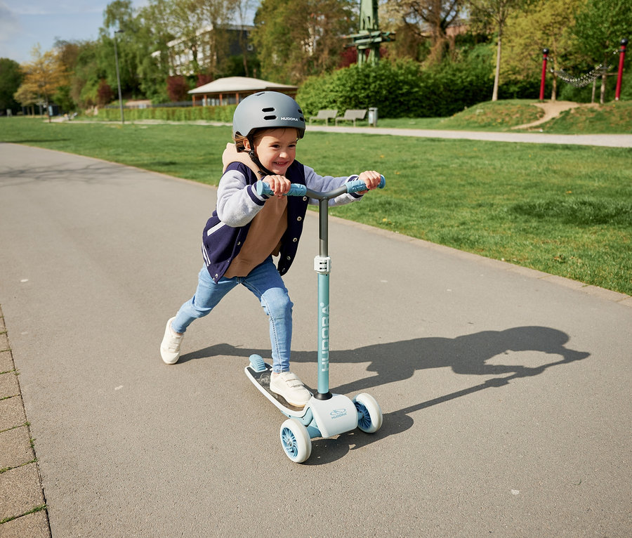 Junge fährt mit blauem HUDORA Tri-Scooter auf Gehweg im Park.