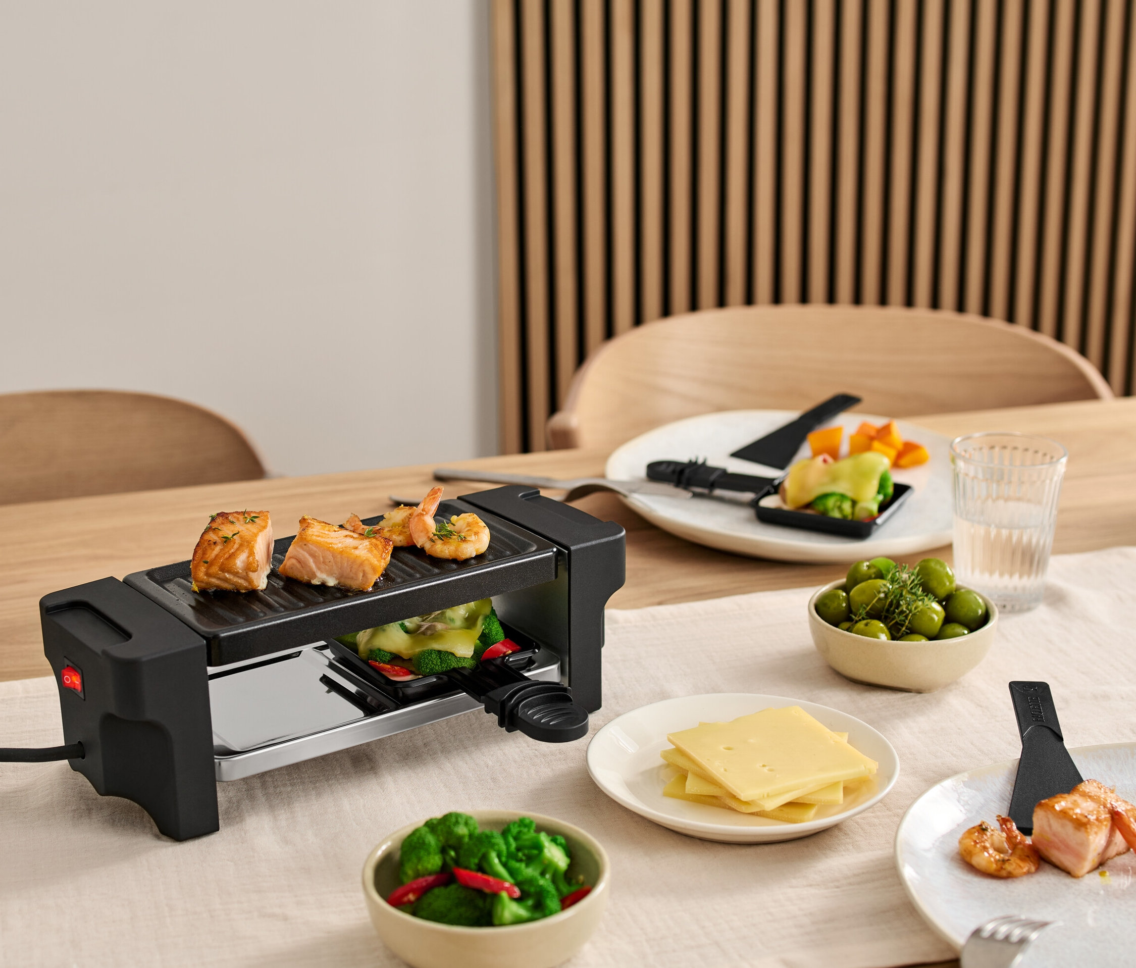 Raclette für 2 steht auf einem Tisch, mit Lachs und Garnelen auf dem Grill. Teller mit Zutaten stehen um den Grill herum.