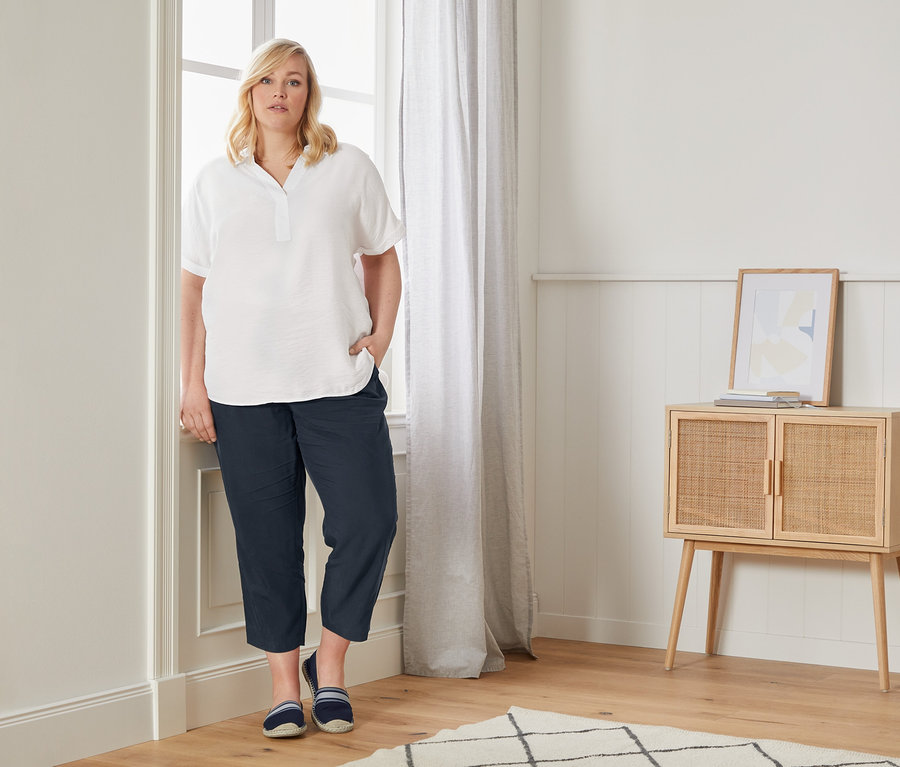 Eine Frau posiert in einer Oversized-Bluse, einer lässigen Chino und Espadrilles am Fenster.