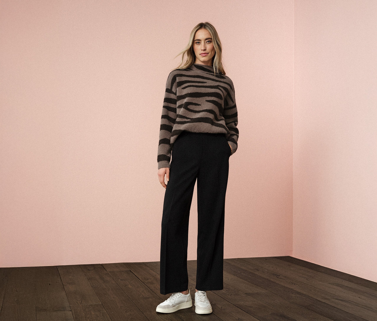 Eine Frau steht vor einer rosa Wand und trägt einen Street One Jacquard-Pullover, eine schwarze Hose und weiße Turnschuhe.