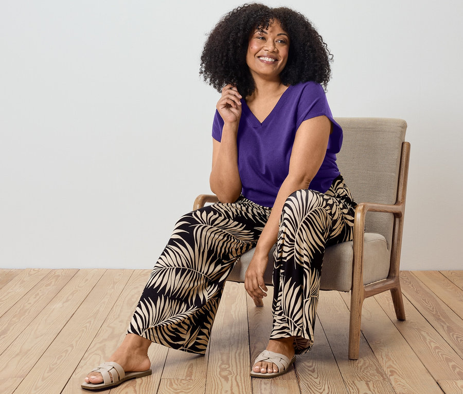 Frau sitzt auf Stuhl und trägt lila Kurzärmliger Feinstrickpullover und bedruckte Palazzohose.