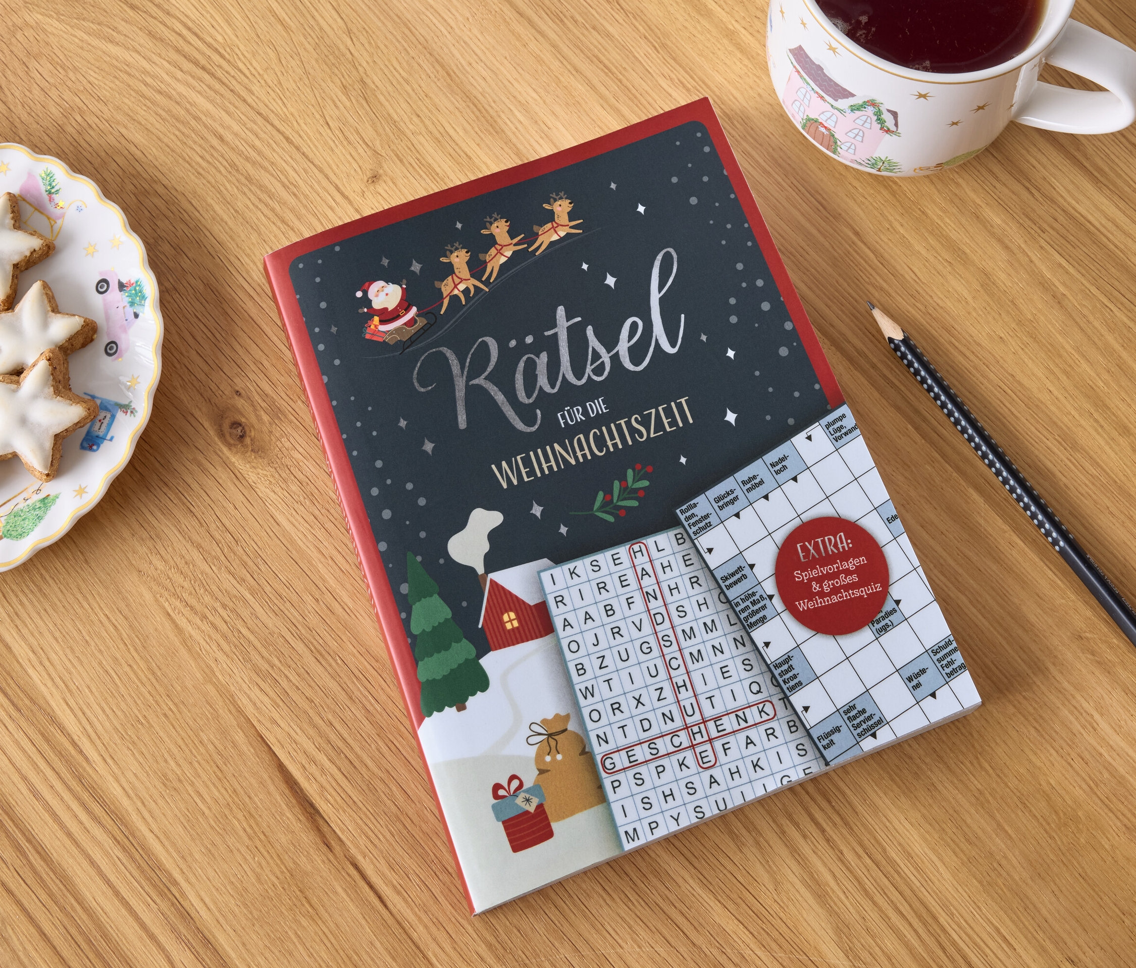 Auf einem Holztisch liegen ein Weihnachtsratselbuch, ein Teller mit sternförmigen Keksen, eine Tasse mit einem Getränk und ein Bleistift.