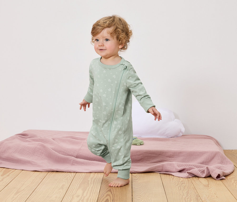 Ein Kleinkind mit lockigen blonden Haaren geht barfuß über einen Holzboden. Es trägt einen hellgrünen Schlafanzug mit Reißverschluss und Herzmuster.