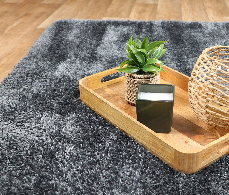 Nahaufnahme eines grauen Teppichs »Silky« mit einer Holzschale, einer Pflanze, einer Kerze und einer Laterne.