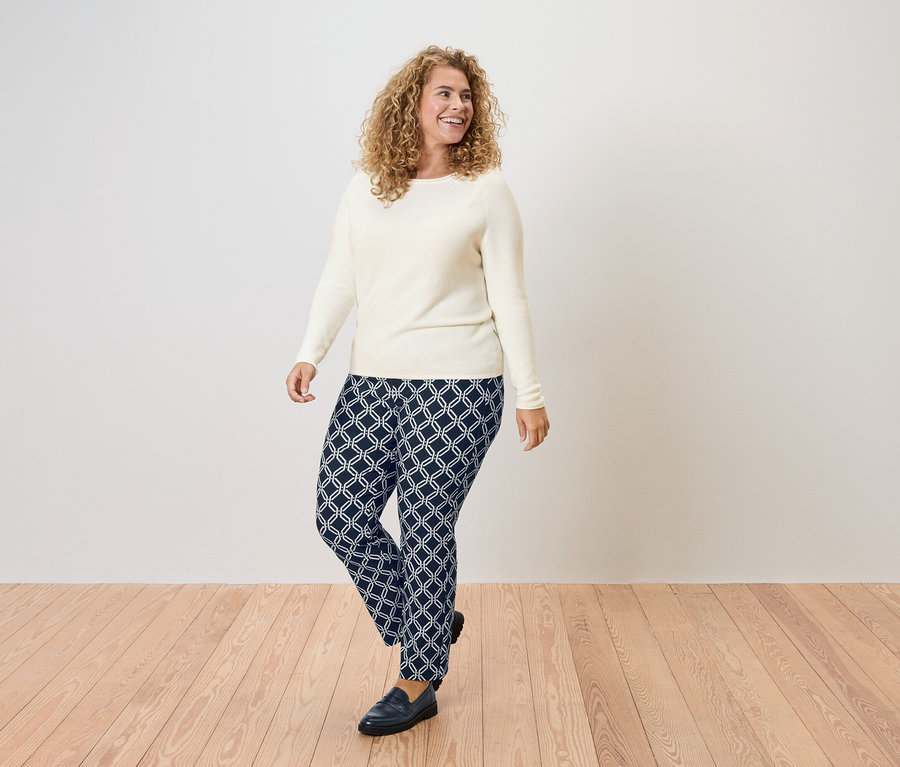Frau mit lockigem Haar, die einen cremefarbenen Pullover und marineblaue Hosen mit geometrischem Muster trägt.