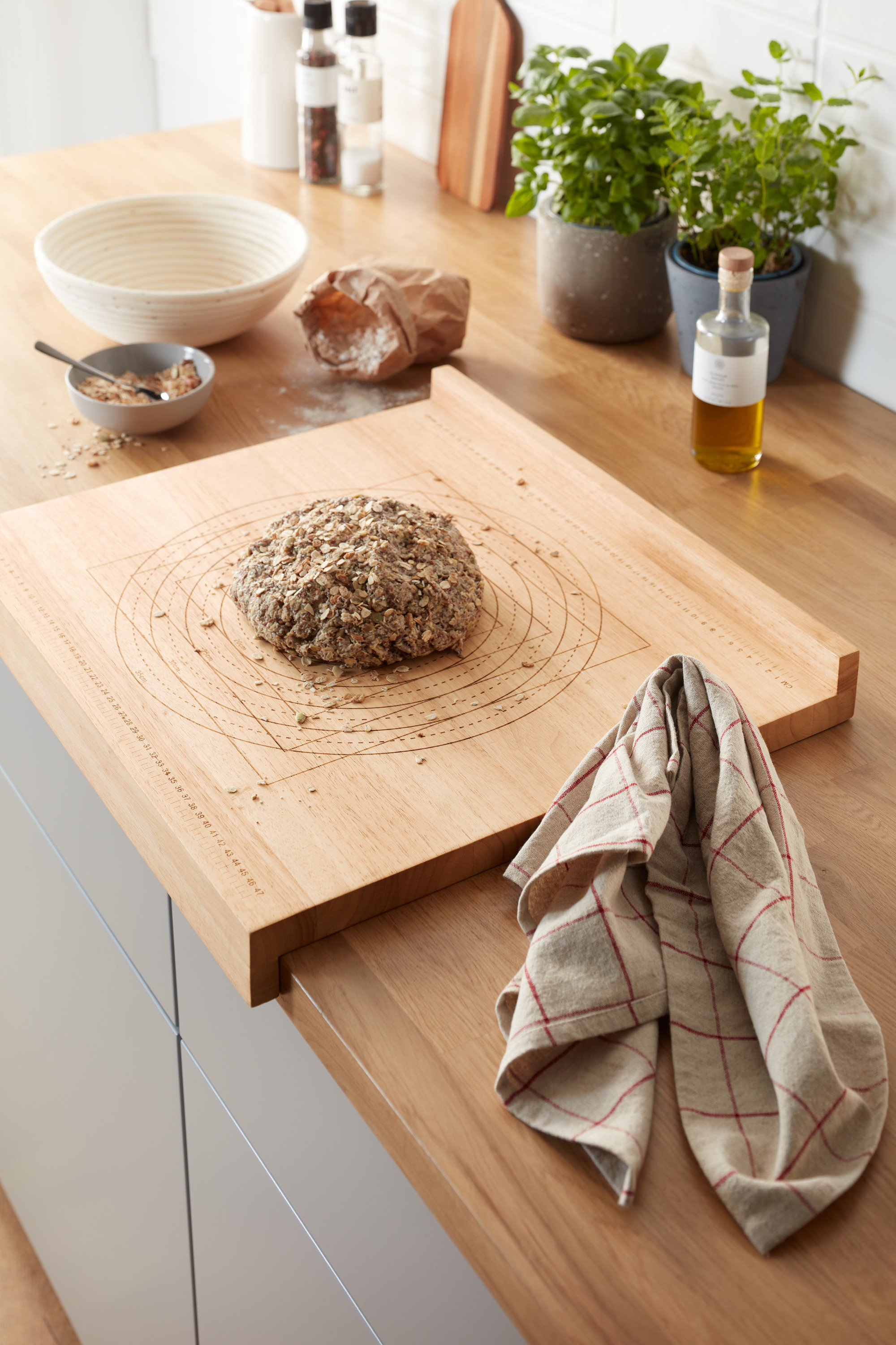 Auf einer Küchentheke liegt ein hölzernes Back- und Arbeitsbrett mit einem Brotlaib. Im Hintergrund sind Kräuter in Töpfen und andere Küchenutensilien zu sehen.