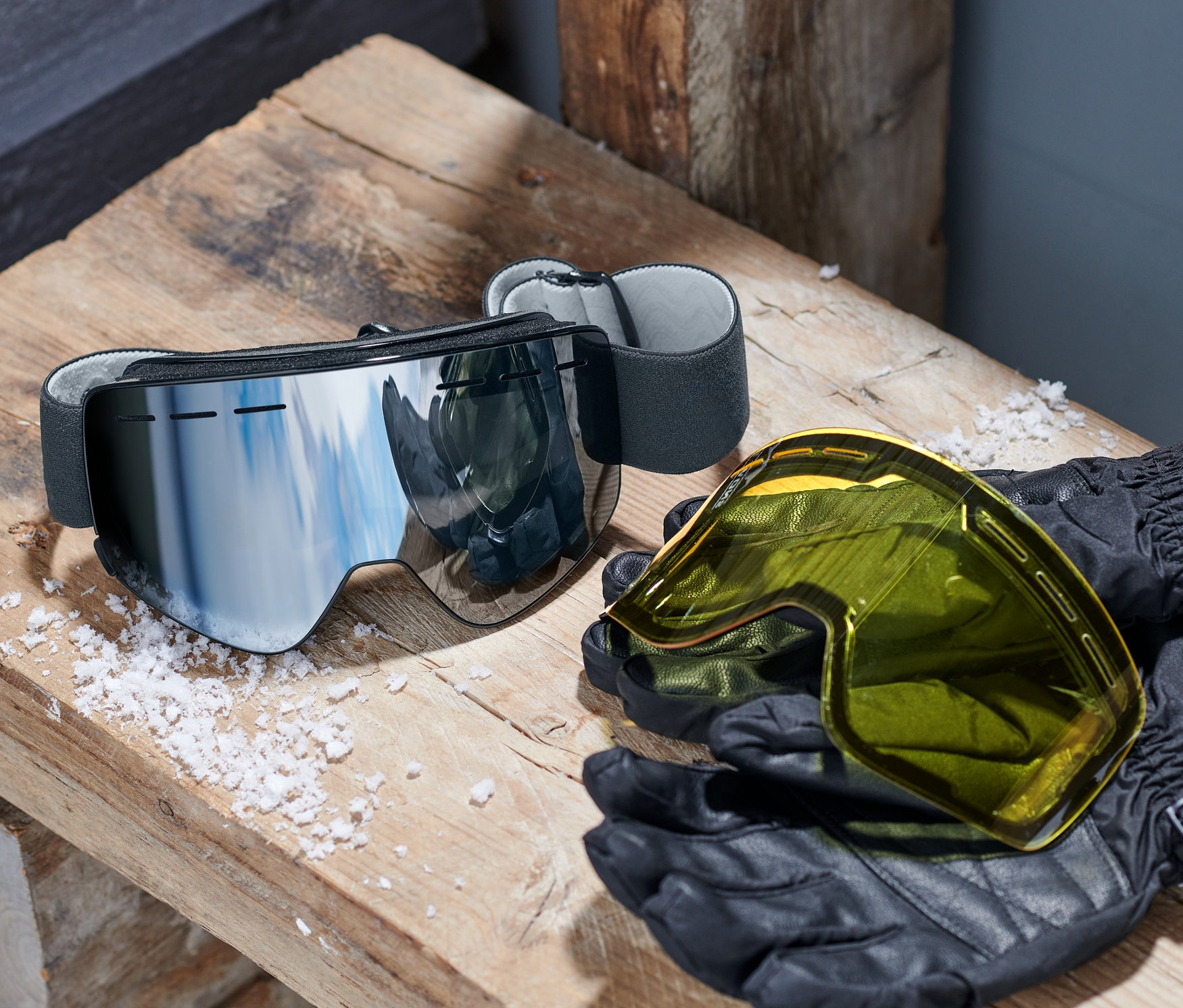 Eine Schneebrille mit magnetischen Wechselscheiben liegt auf einer Holzbank neben einem gelben Visier und schwarzen Handschuhen.