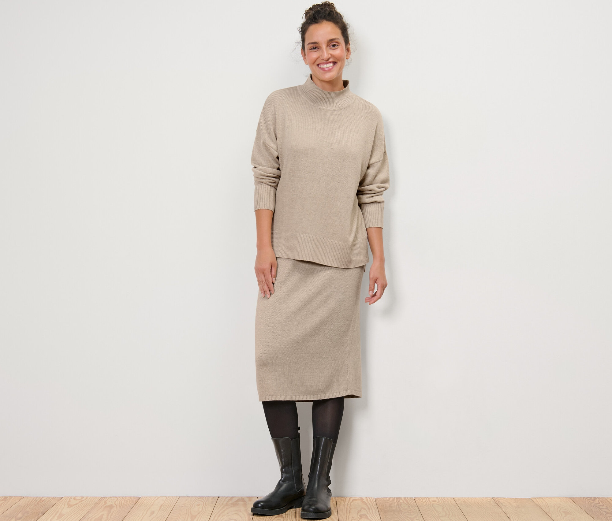 Eine Frau steht und trägt einen Feinstrickpullover mit Stehkragen und einen Strickrock.