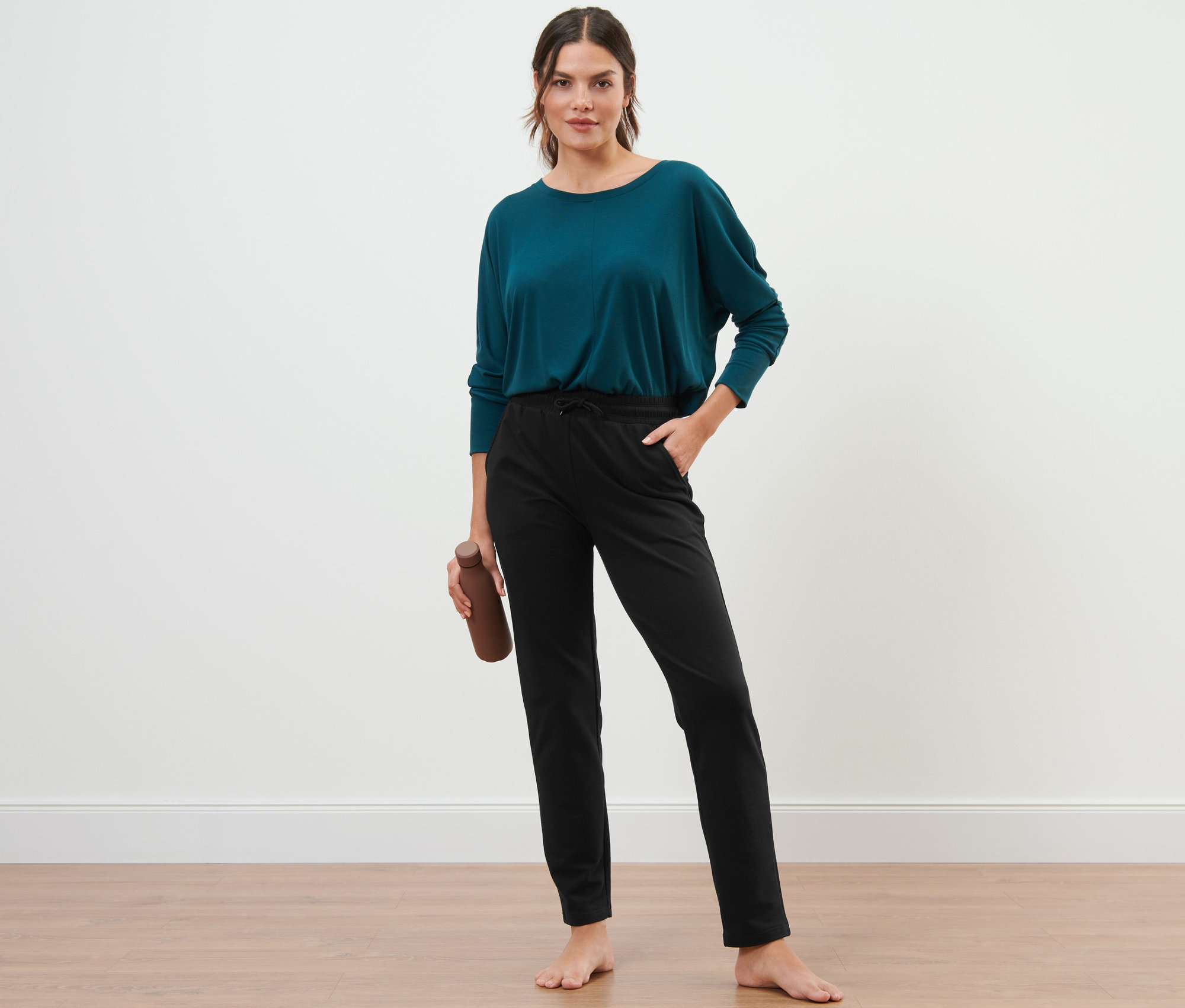 Eine barfüßige Frau steht, hält eine braune Wasserflasche und trägt eine schwarze Sweathose.