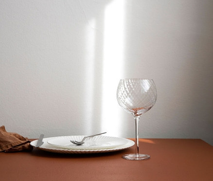 Auf einem Tisch liegen ein Teller, Besteck, ein braunes Tuch und 6 Weingläser.