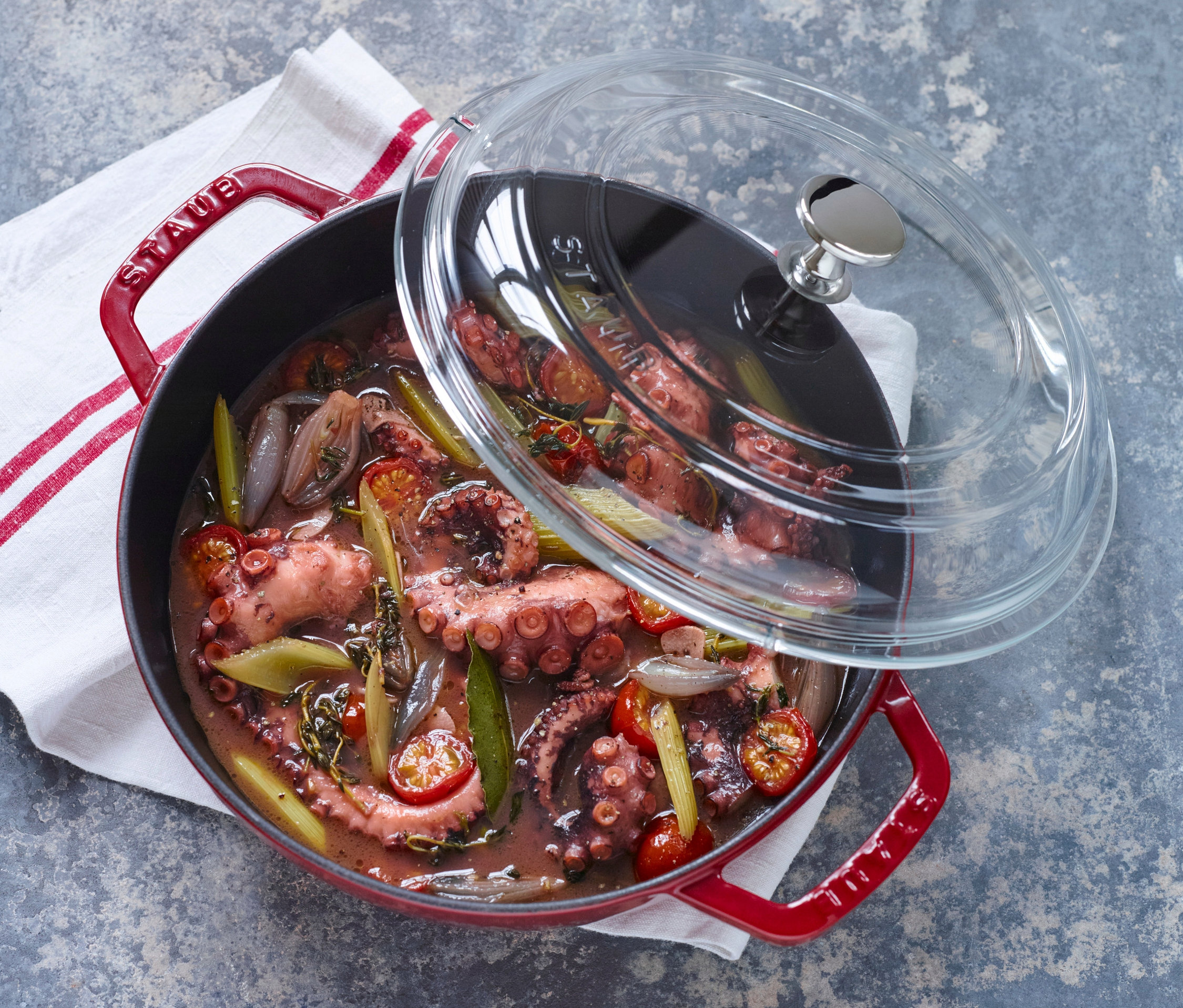 Ein roter STAUB Gusseisen-Bräter »Braisers« mit Glasdeckel, gefüllt mit gekochtem Gemüse und Oktopus, steht auf einer grauen Oberfläche neben einem weißen Geschirrtuch.