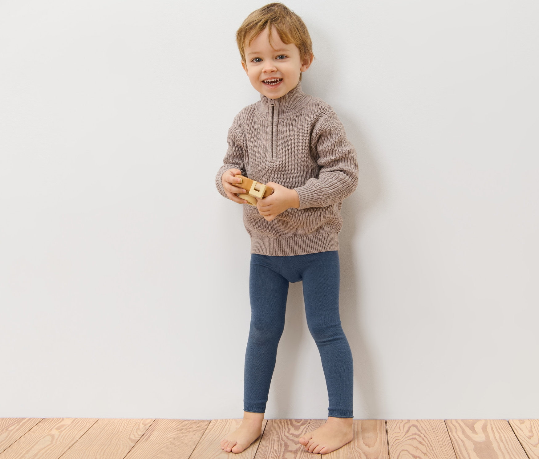 Ein Junge steht an einer Wand und spielt mit einem Holzspielzeug. Er trägt blaue Strickleggings und einen braunen Pullover. 3 Kinder-Strickleggings.