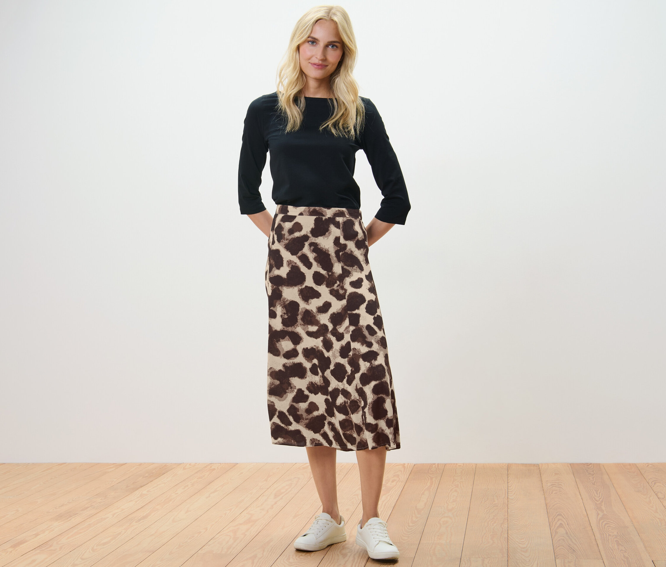 Blonde Frau steht mit Händen hinter dem Rücken, trägt eine schwarze Bluse und einen bedruckten Midi-Rock.