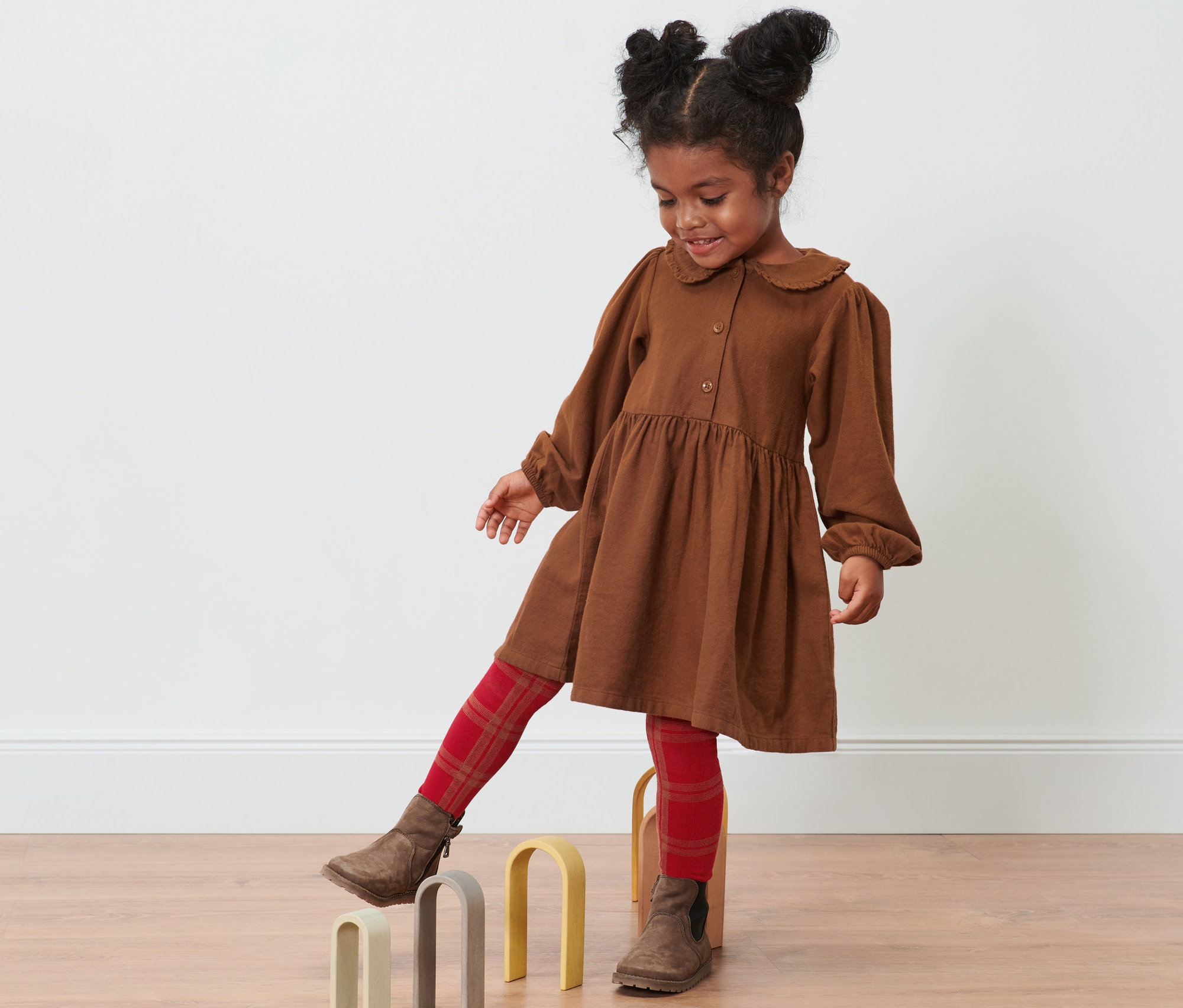 Mädchen mit Duttfrisur im cognacfarbenen Webkleid balanciert über Holzbögen.
