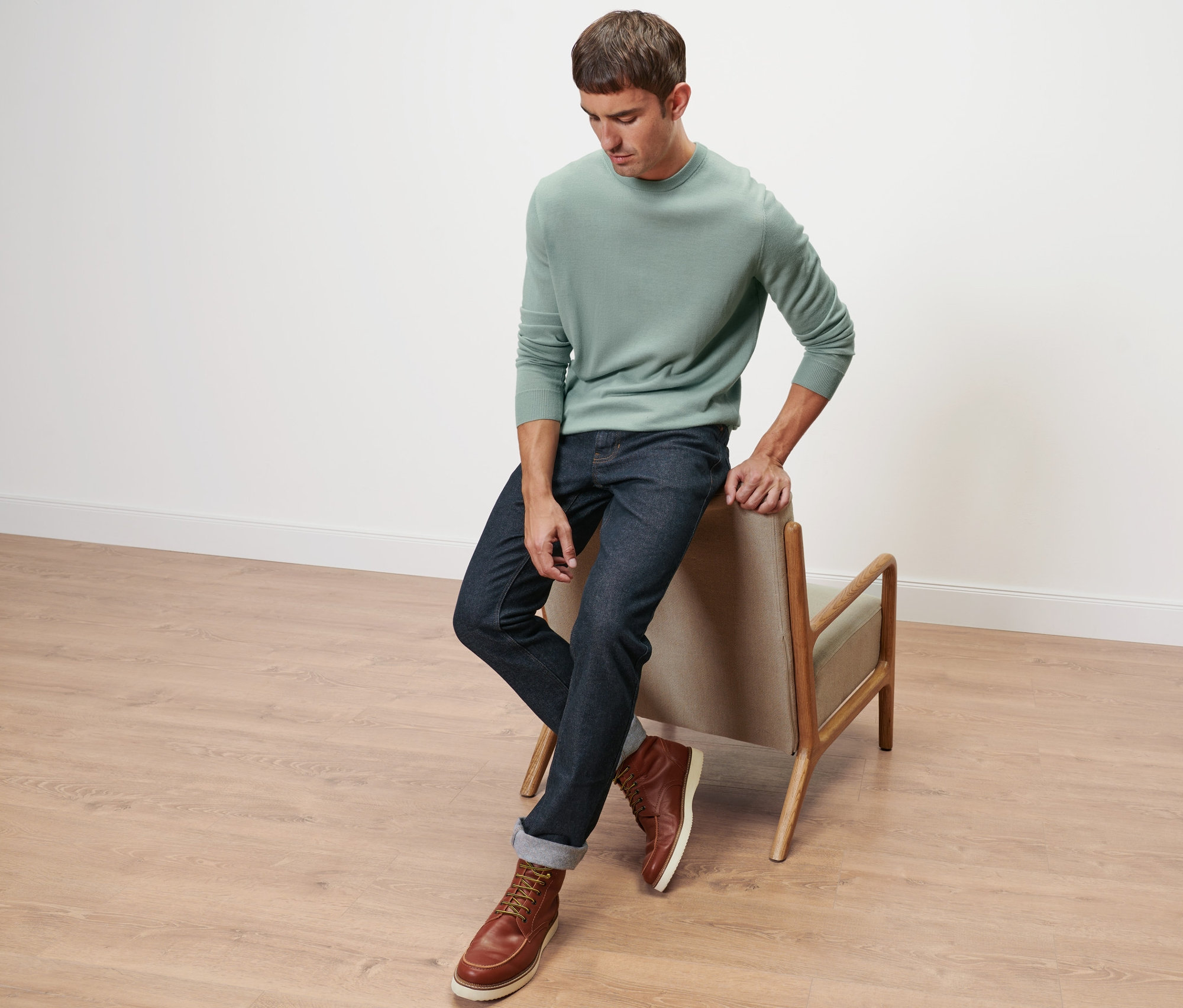 Mann sitzt auf einer Stuhllehne, trägt einen hellgrünen Merino-Pullover und Jeans.