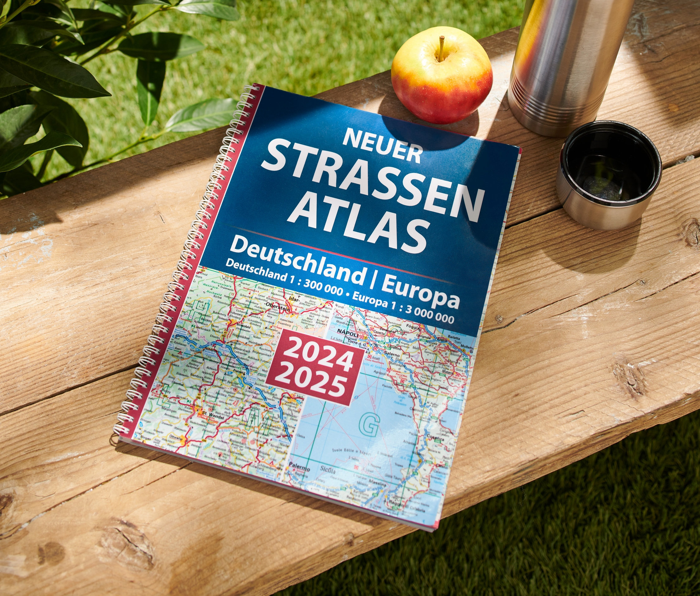 Auf einem Holztisch liegen ein Apfel, eine Thermoskanne und das Buch: Neuer Straßen-Atlas Deutschland/Europa 2024/2025.