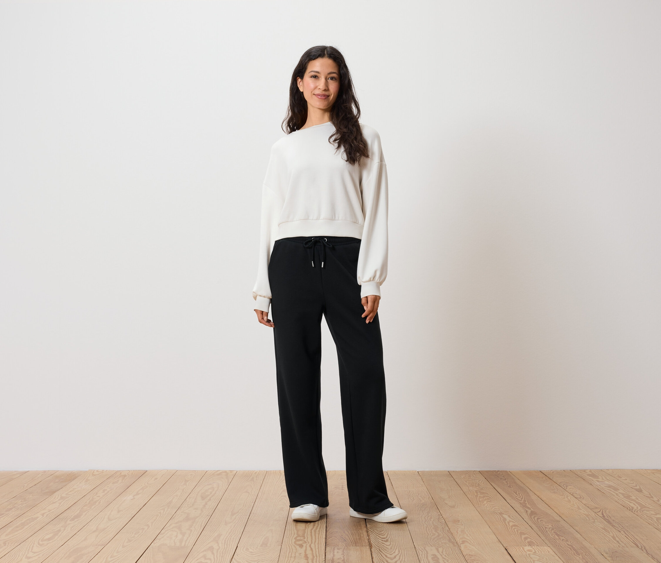 Eine Frau steht auf einem Holzboden und trägt ein weißes Sweatshirt und eine schwarze Hose.