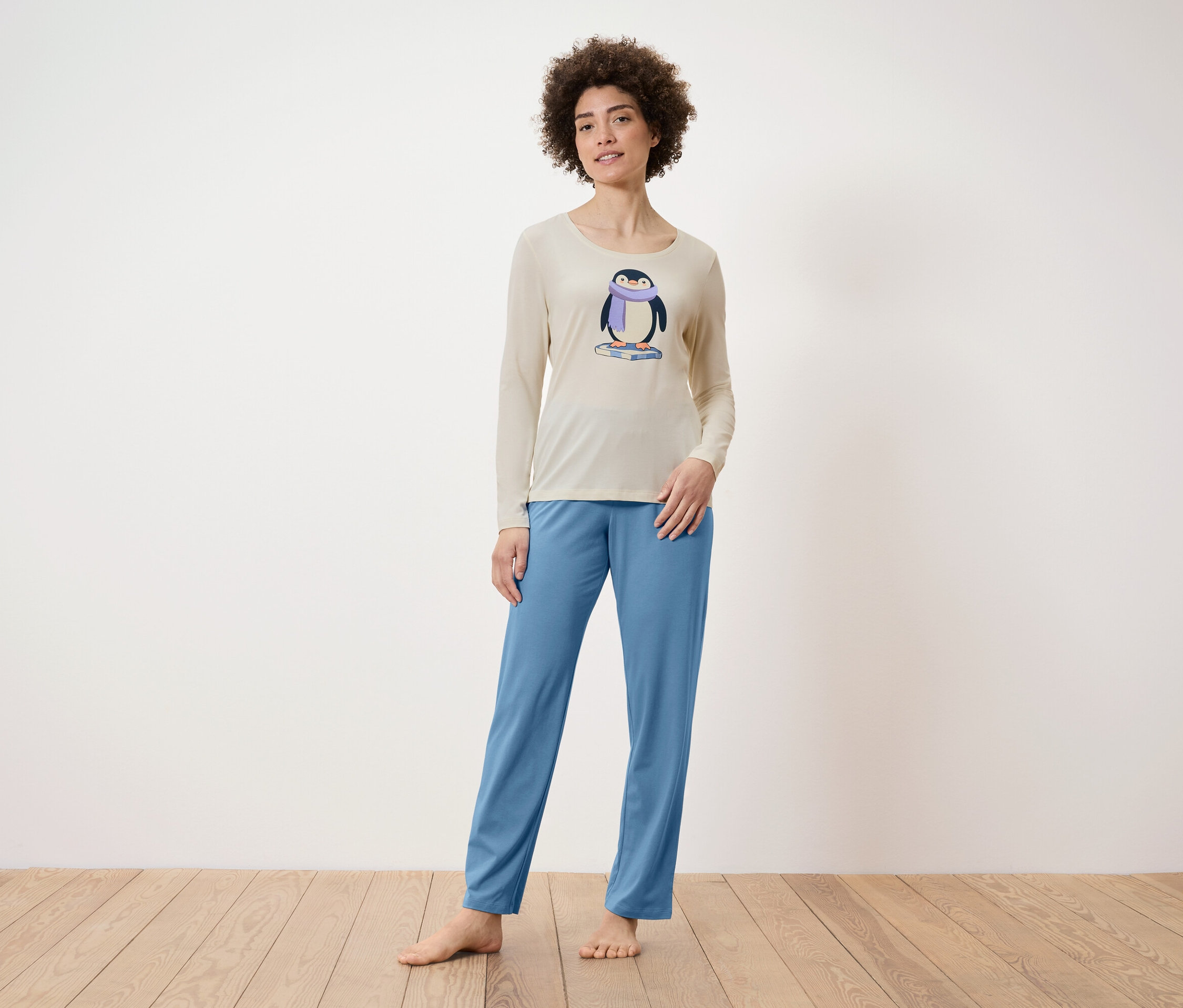 Frau steht barfuß und trägt beigen Pyjamaoberteil mit Pinguin und blaue Pyjamahose.