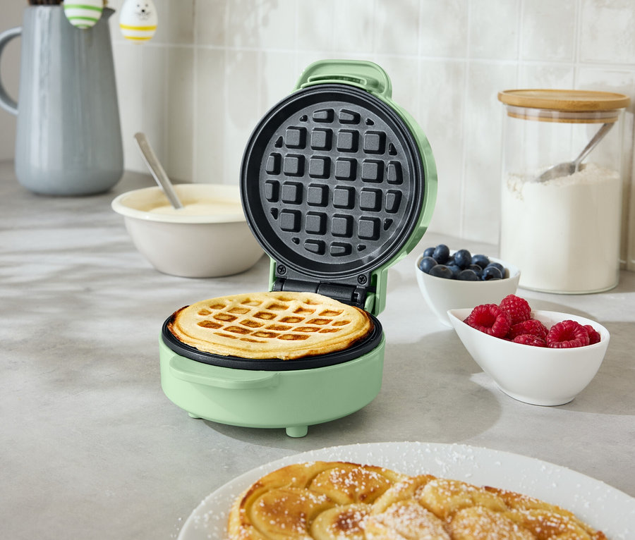 Ein geöffnetes, rundes, mintgrünes Waffeleisen mit einer Waffel darin steht auf einer Küchentheke. Daneben befinden sich zwei spiralförmige Backwaren auf einem Teller, eine Schüssel mit Blaubeeren und eine Schüssel mit Himbeeren.