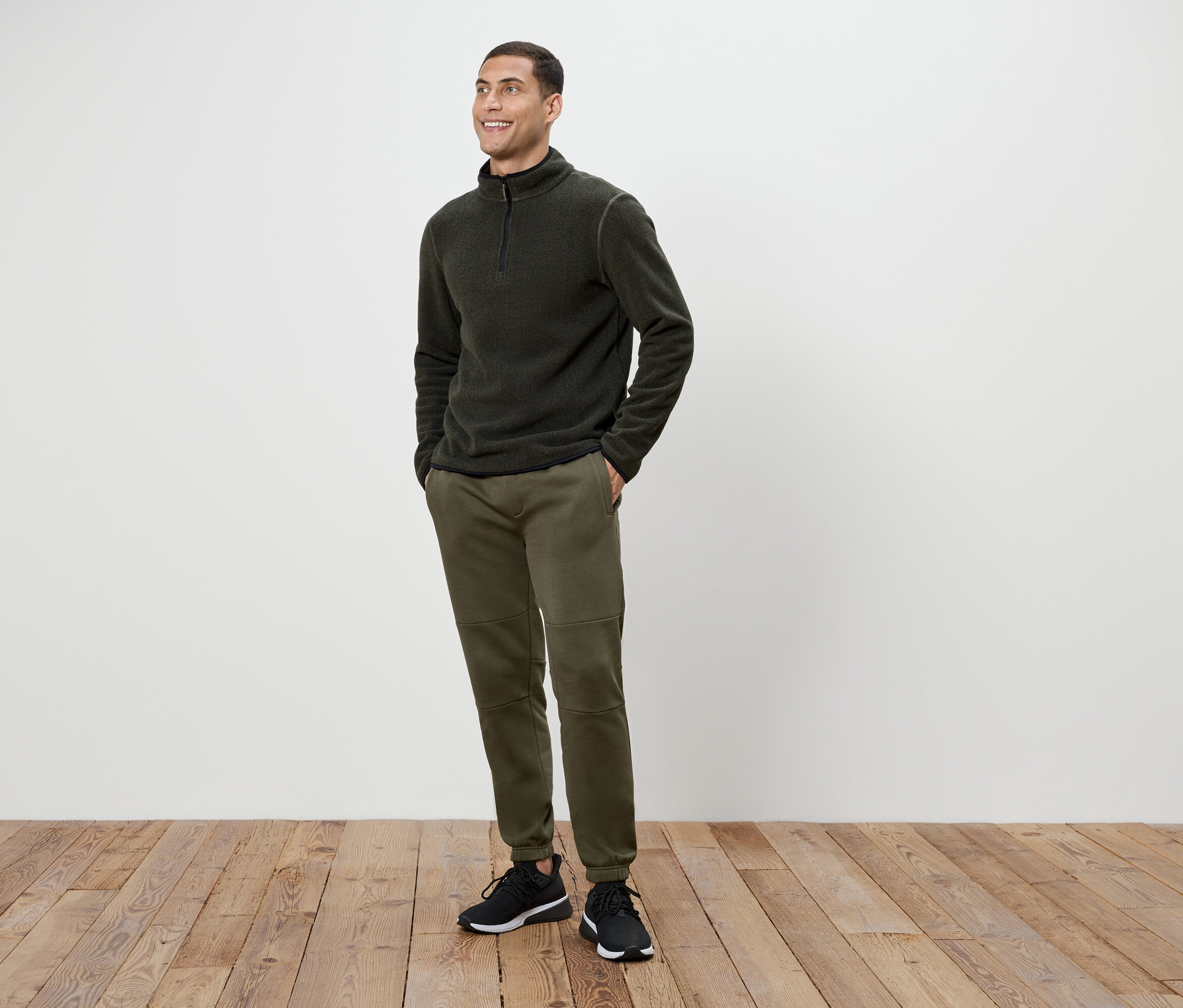 Ein Mann in olivgrünen Hosen und einem dunkelgrauen Fleece-Pullover mit Reißverschluss steht auf einem Holzboden.