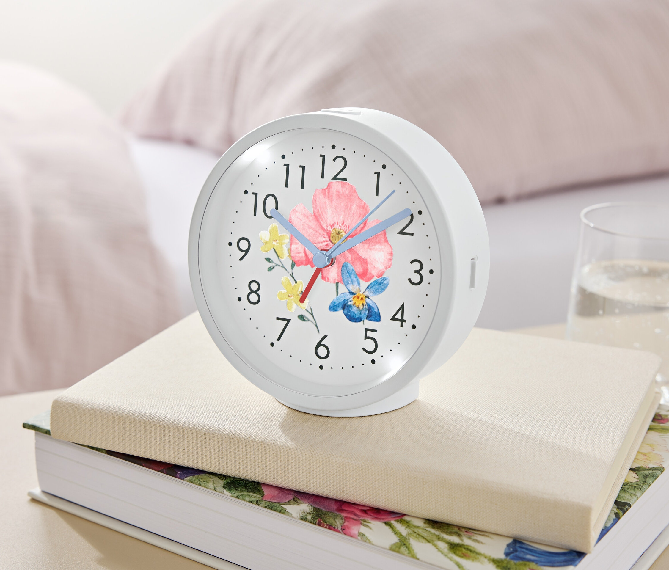 Ein weißer Wecker mit Blumenmuster liegt auf Büchern neben einem Glas Wasser.