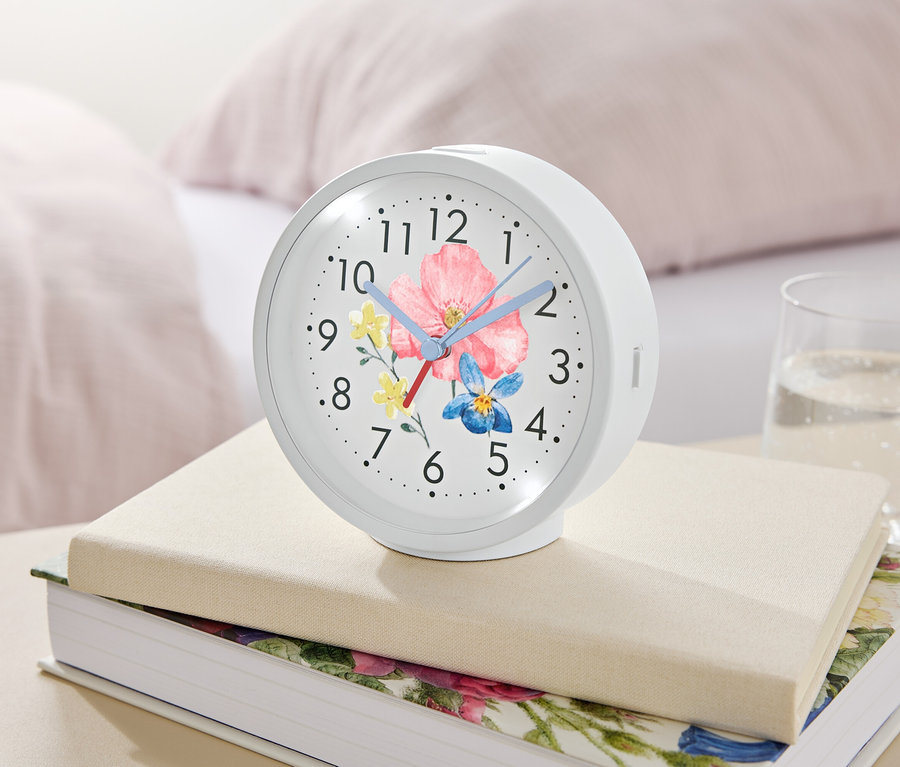 Ein weißer Wecker mit Blumenmuster liegt auf Büchern neben einem Glas Wasser.