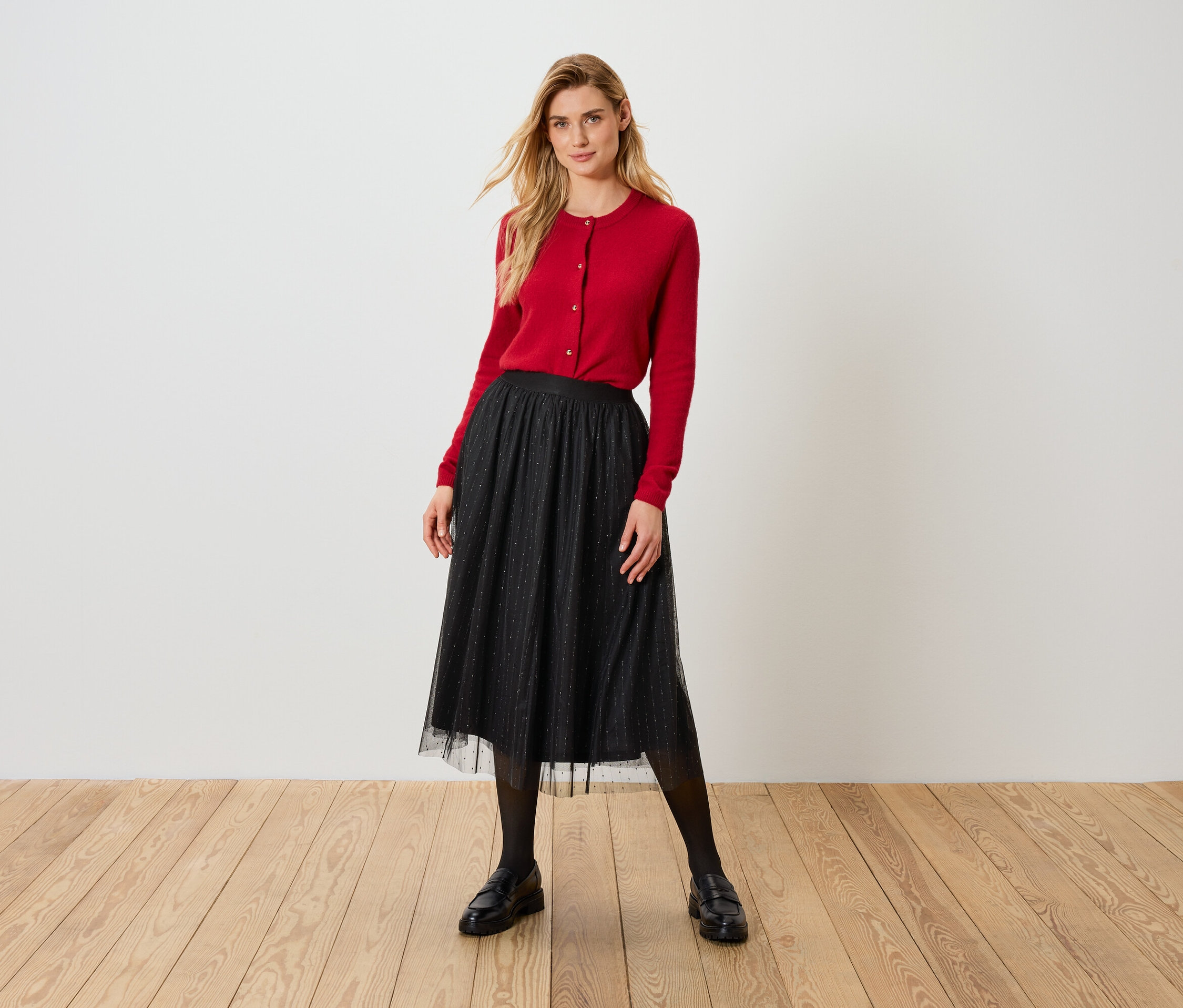 Blonde Frau in roter Strickjacke und schwarzem Tüllrock steht auf Holzboden.