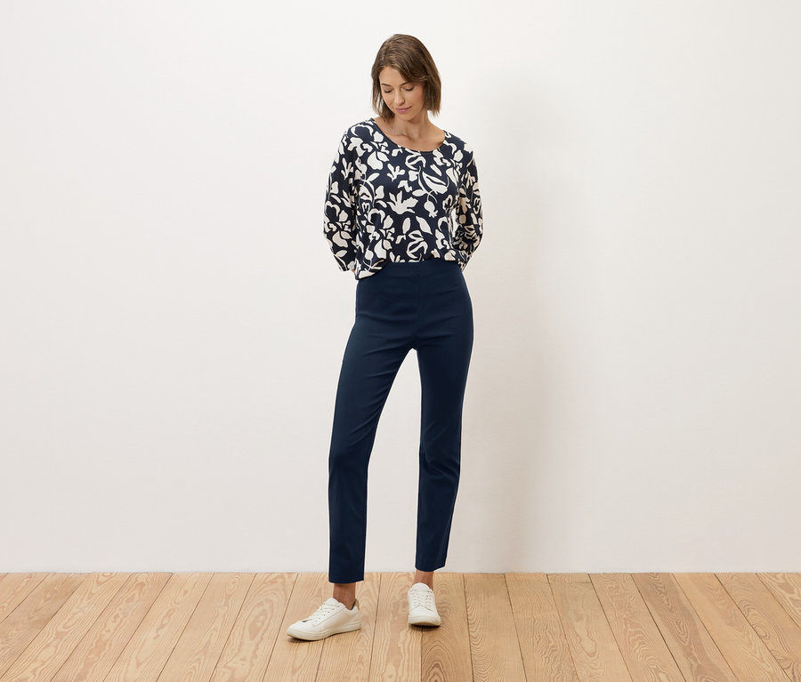 Eine Frau steht vor einer weißen Wand, gekleidet in eine marineblaue Hose und ein schwarz-weiß gemustertes Oberteil.