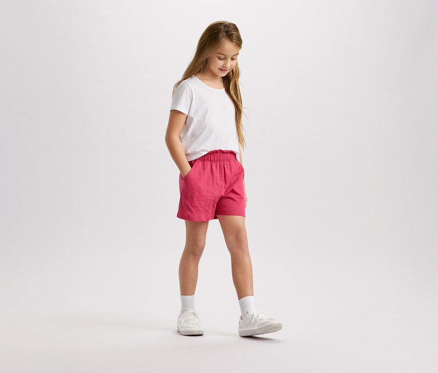 Mädchen mit blonden Haaren, weißem T-Shirt und pinkfarbenen Shorts steht da und schaut nach unten.