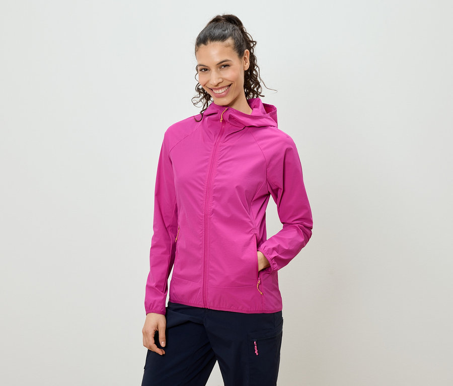 Frau mit hochgesteckten Haaren, die eine pinkfarbene Jacke mit Kapuze und marineblaue Hosen trägt und in die Kamera lächelt.