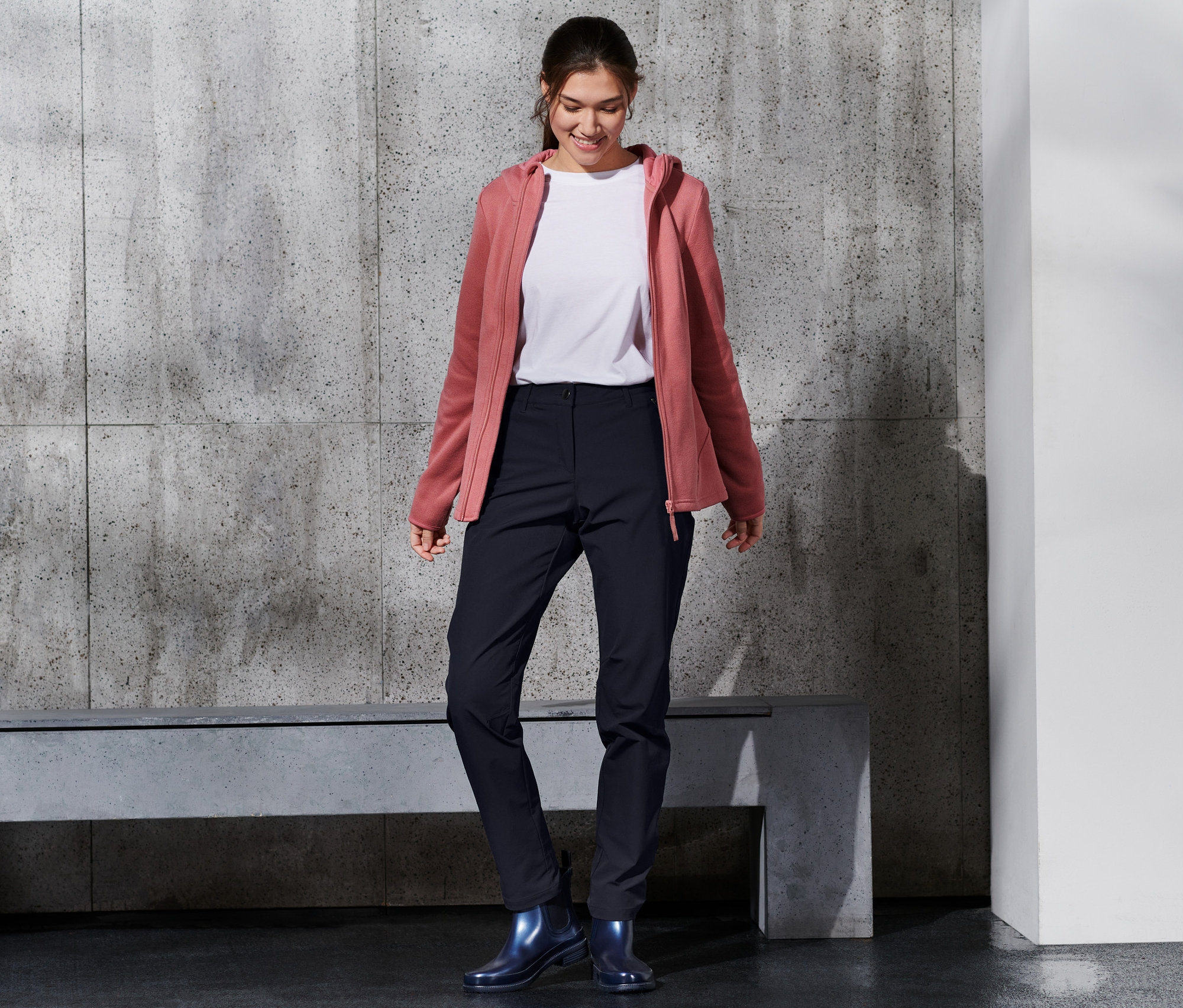 Lächelnde Frau trägt eine Softshell-Hose, ein weißes T-Shirt und eine rosafarbene Kapuzenjacke.